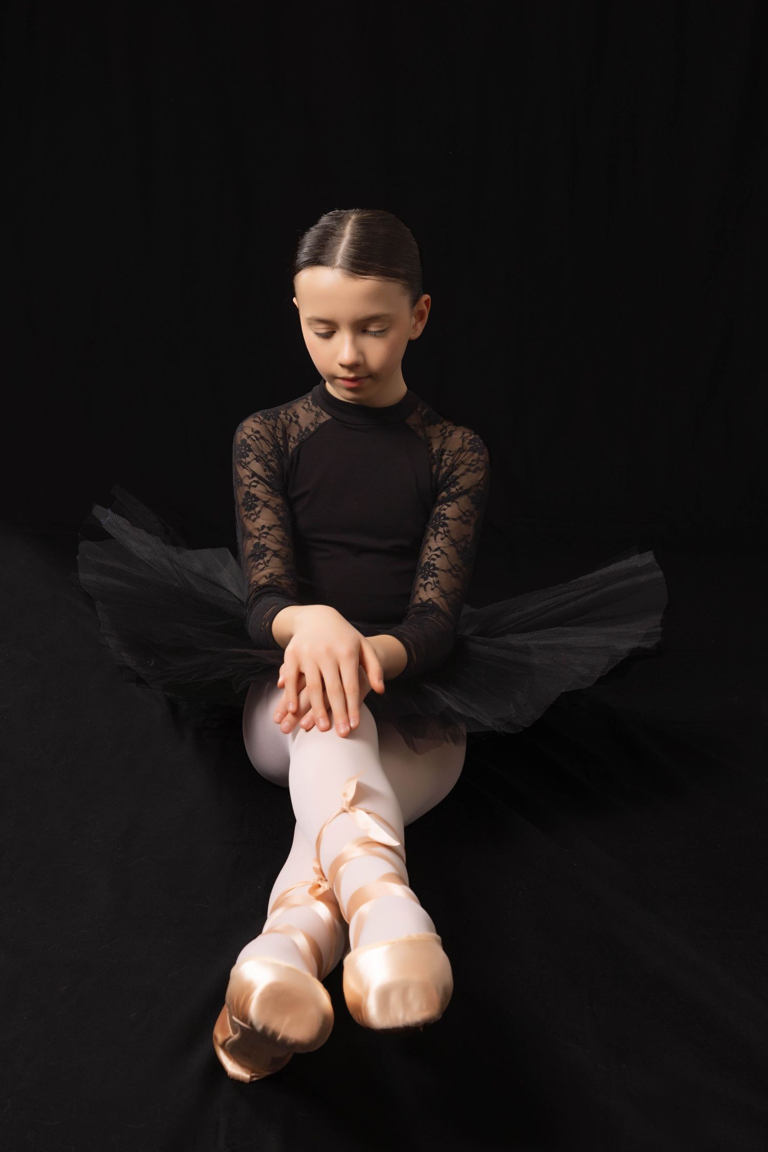 Portrait of a ballet dancer in black leotard and lace sleeves adjusts pink pointe shoes against dark background.