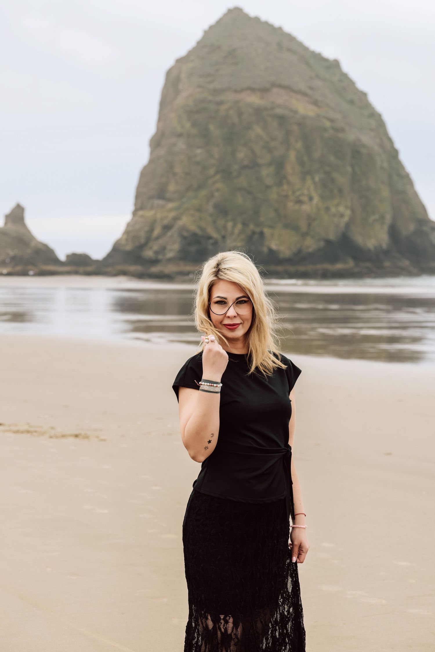 Blonde woman in black lace dress posing on sandy beach with iconic Haystack Rock in background.