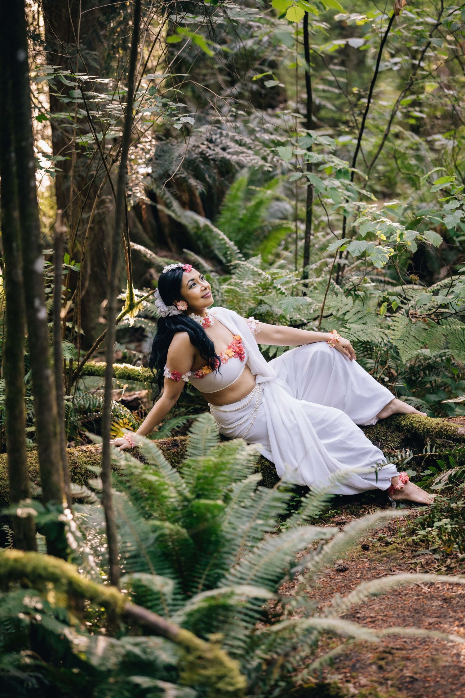 Woman in white dress and floral top poses gracefully among ferns and trees in a lush forest setting.