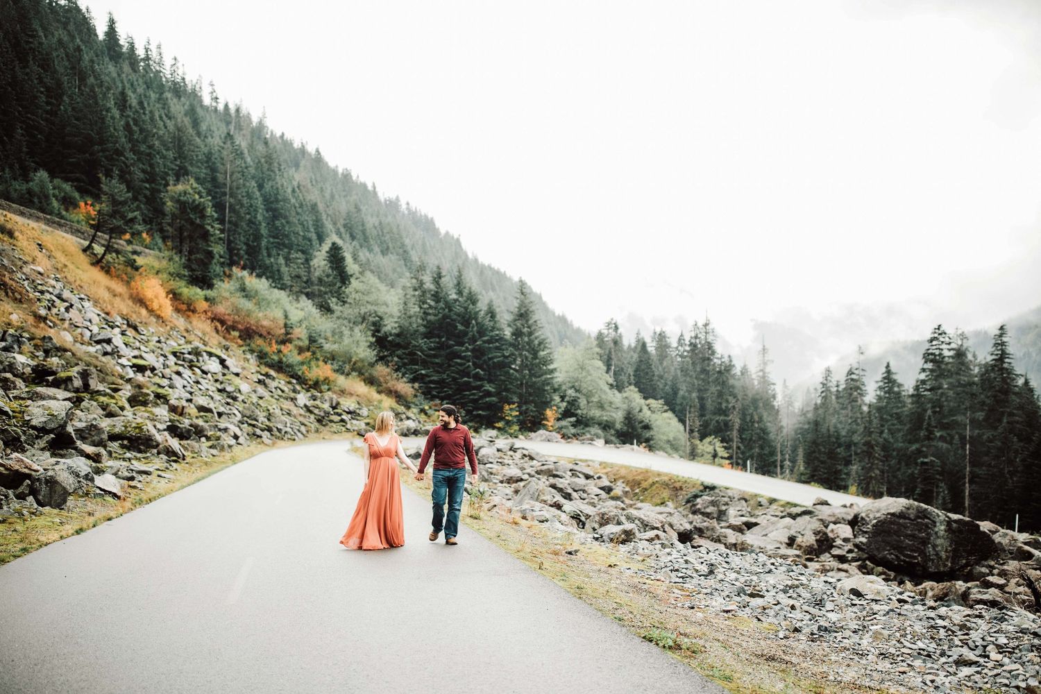 Couple walking hand in hand on a mountain road surrounded by tall pine trees and misty peaks, woman in flowing orange dress.