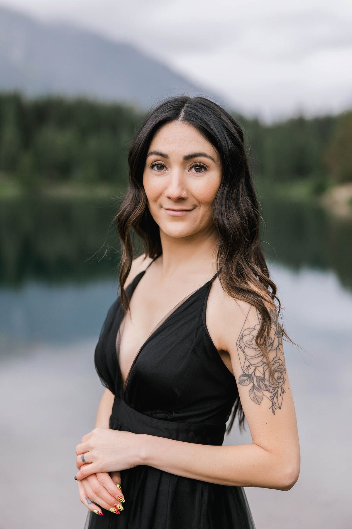 Portrait of a girl in black dress poses elegantly against scenic mountain lake backdrop.