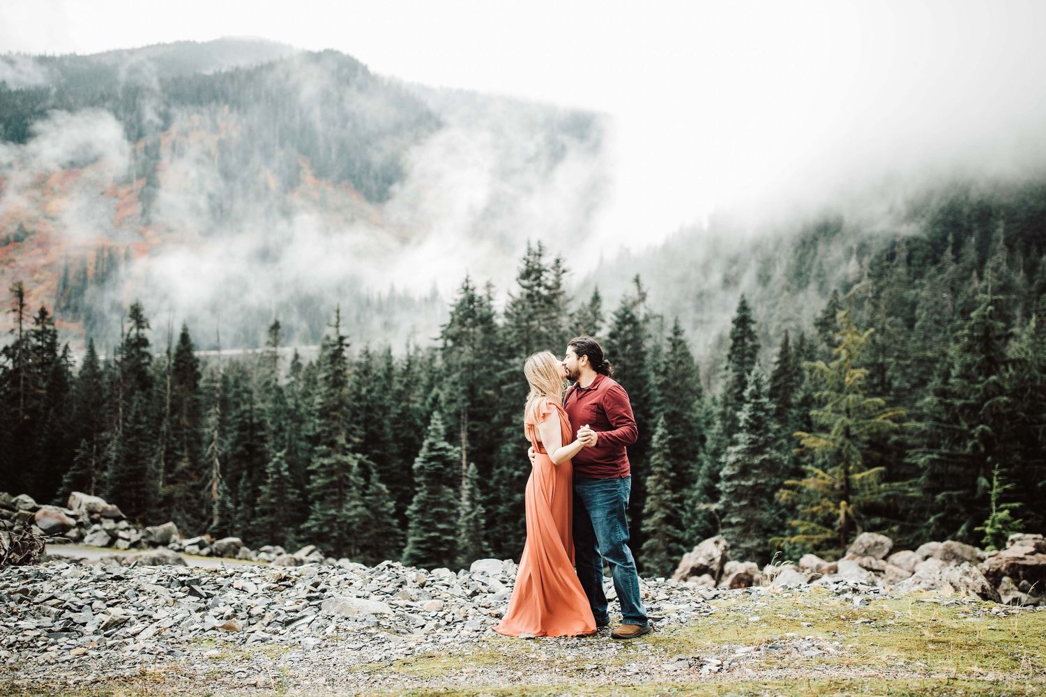 Couple embracing in a romantic outdoor engagement photo with misty mountains and evergreen forest backdrop.