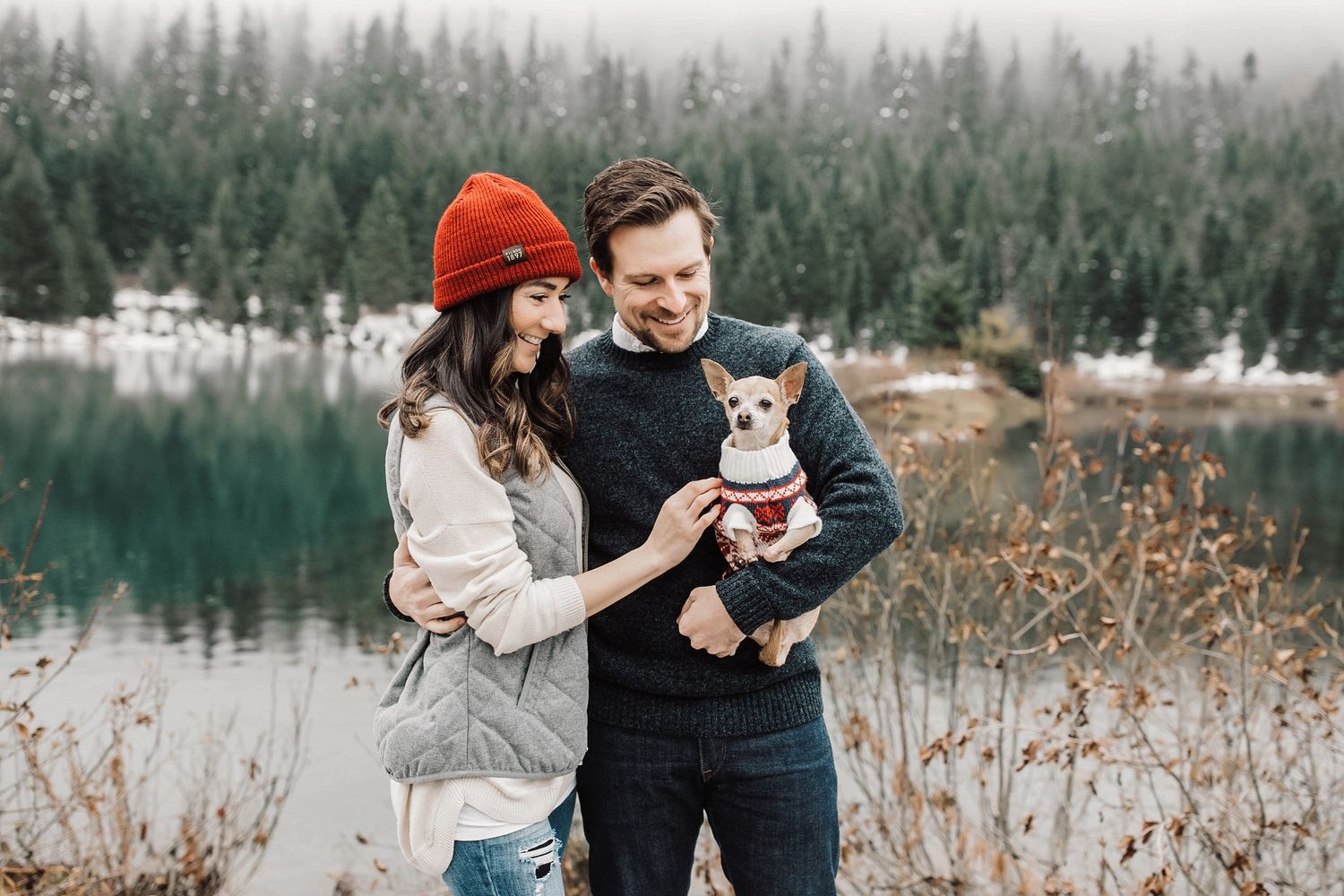 Couple smiling with small dog in sweater by snowy lake and pine forest in winter.
