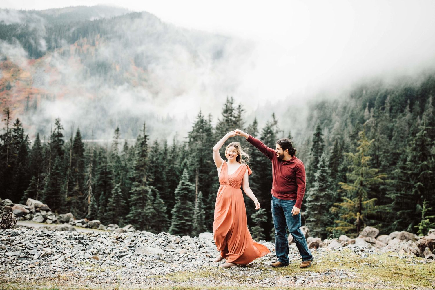 Couple dancing outdoors with misty forest and mountains in background, woman in flowing orange dress.
