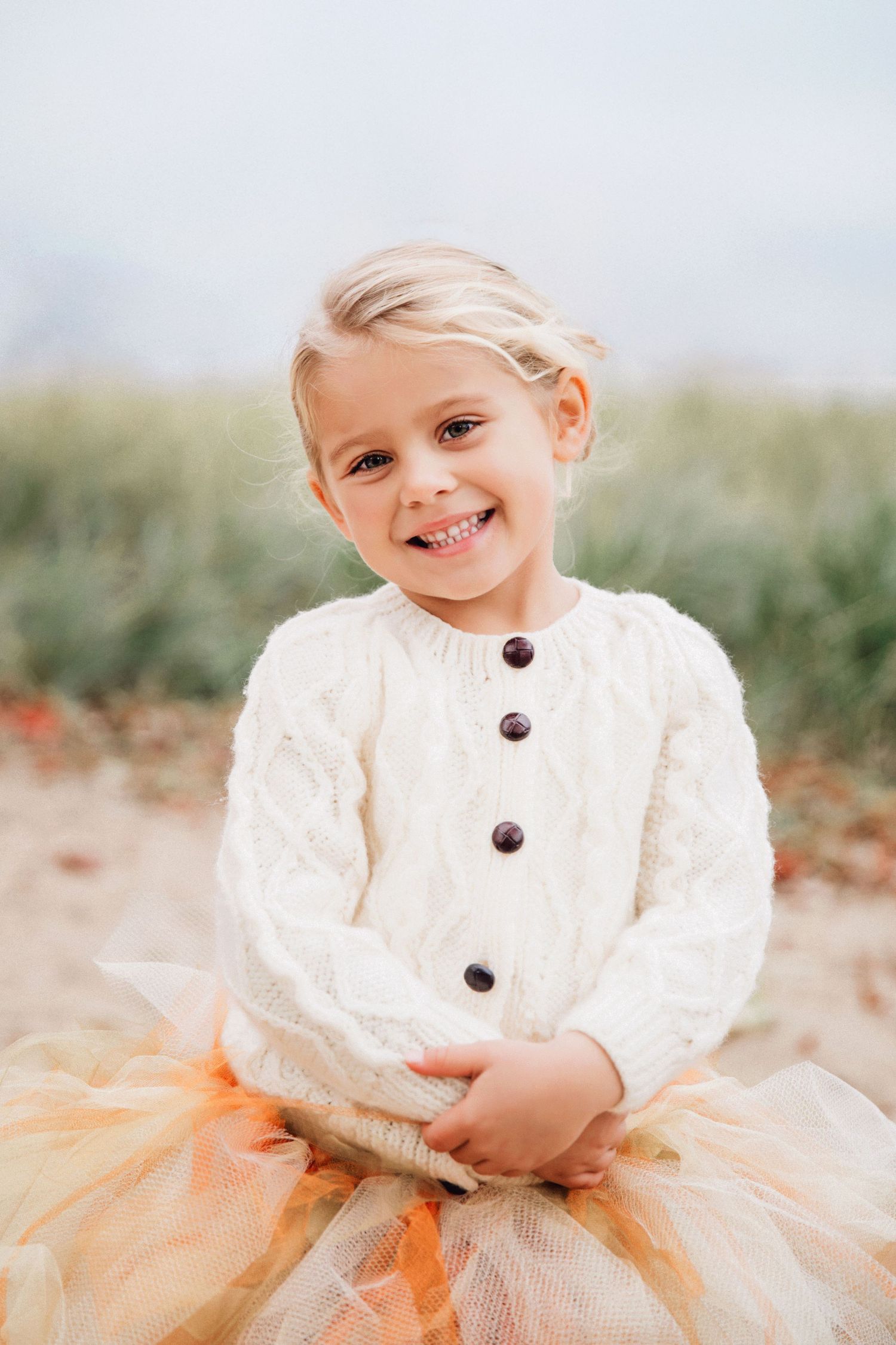 A photo of a child in Seattle in a white knit sweater and orange tulle skirt smiles while sitting outdoors in soft natural lighting.