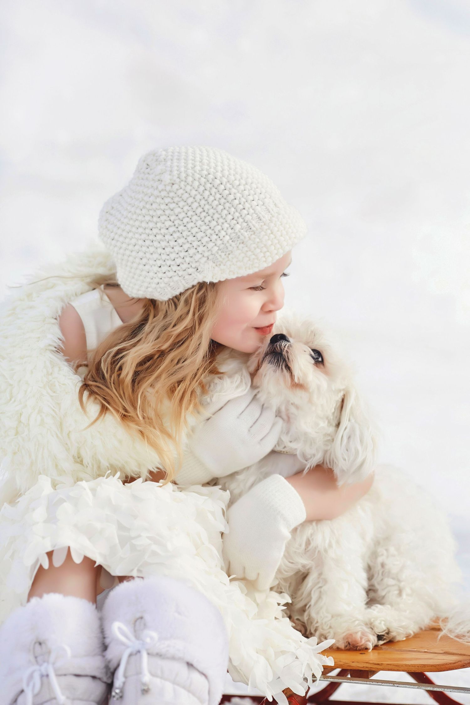 A portrait of a child in a white knit hat and winter clothing snuggles with her fluffy white dog in a winter wonderland setting.