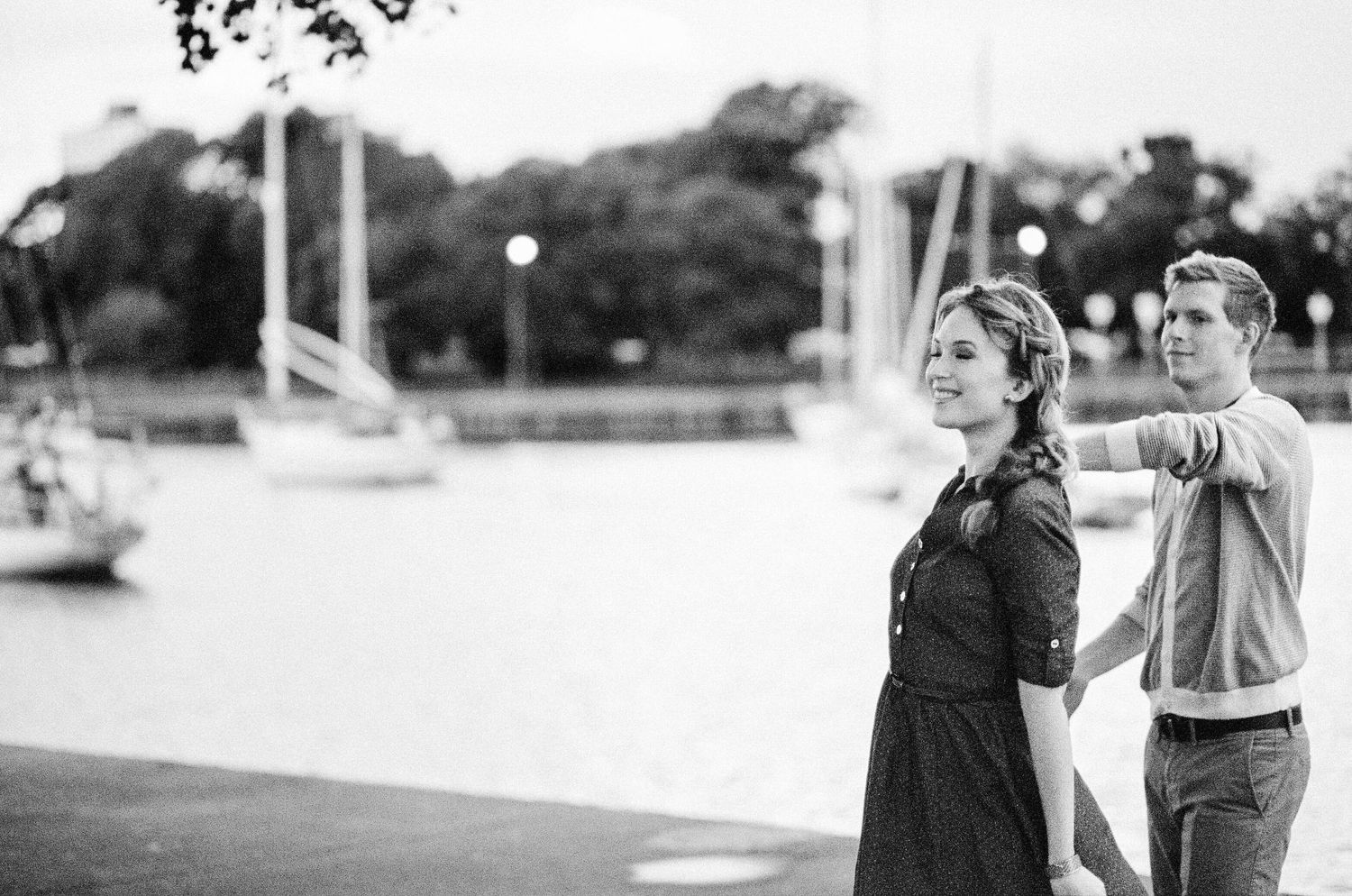 Black and white photo of a young couple holding hands near a marina with sailboats in the background.