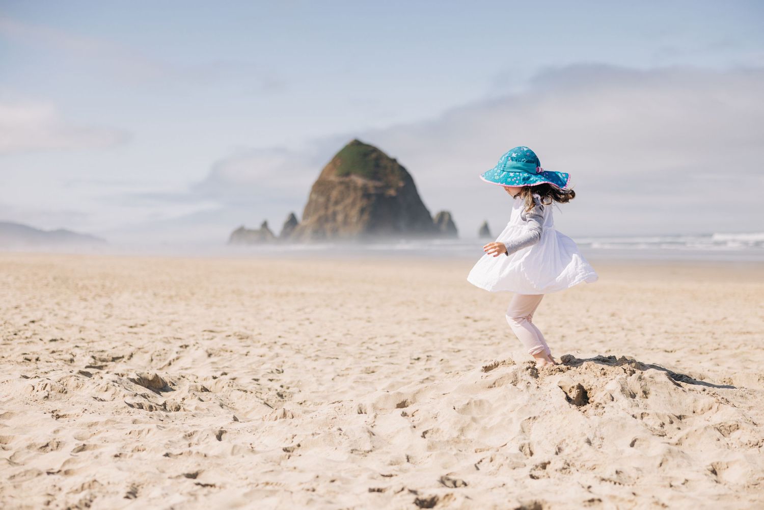 A young girl in a white dress and blue sun hat plays on a sandy beach with Haystack Rock in the background.