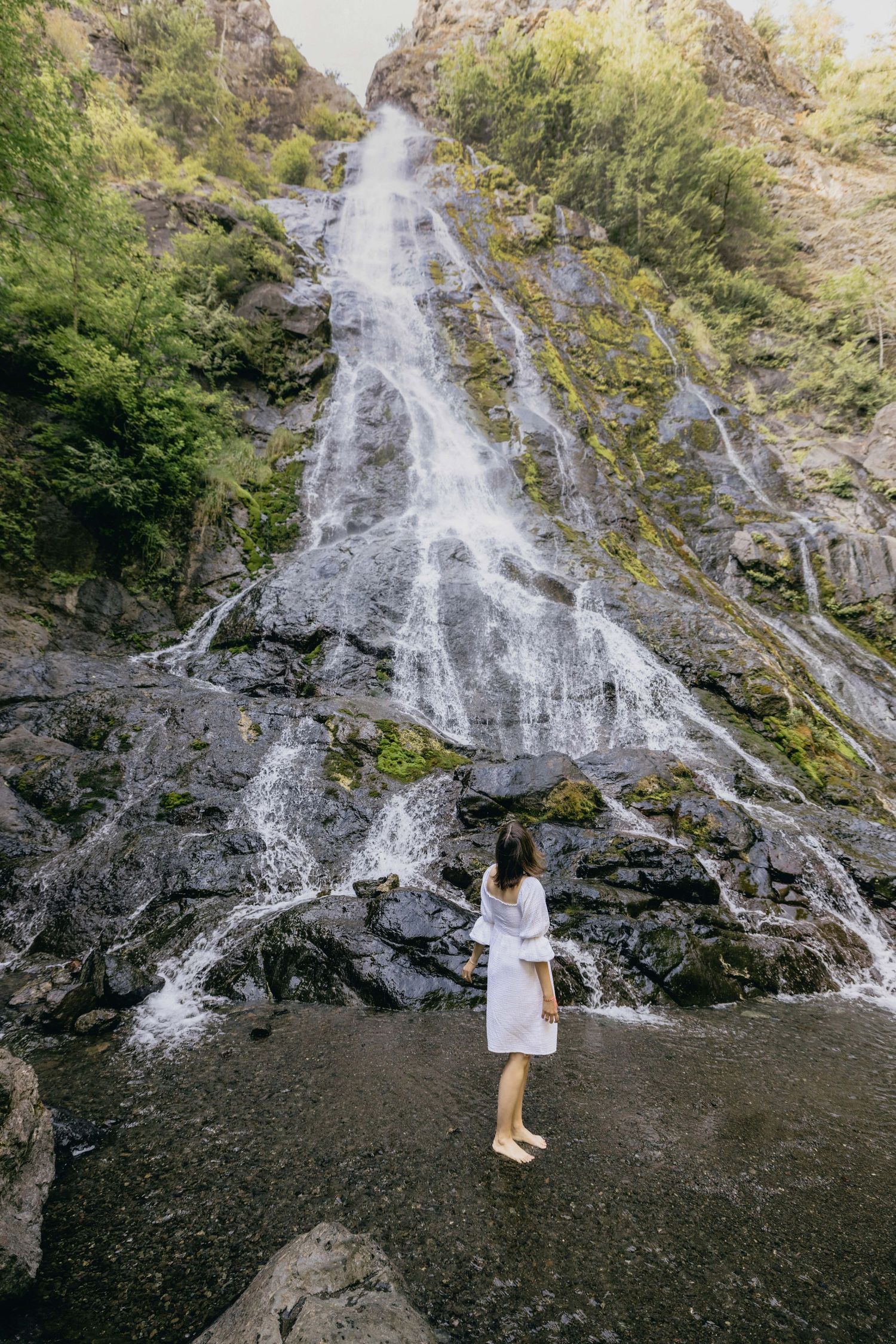 Woman in white dress standing before a tall cascading waterfall surrounded by lush green trees and rocky cliffs.
