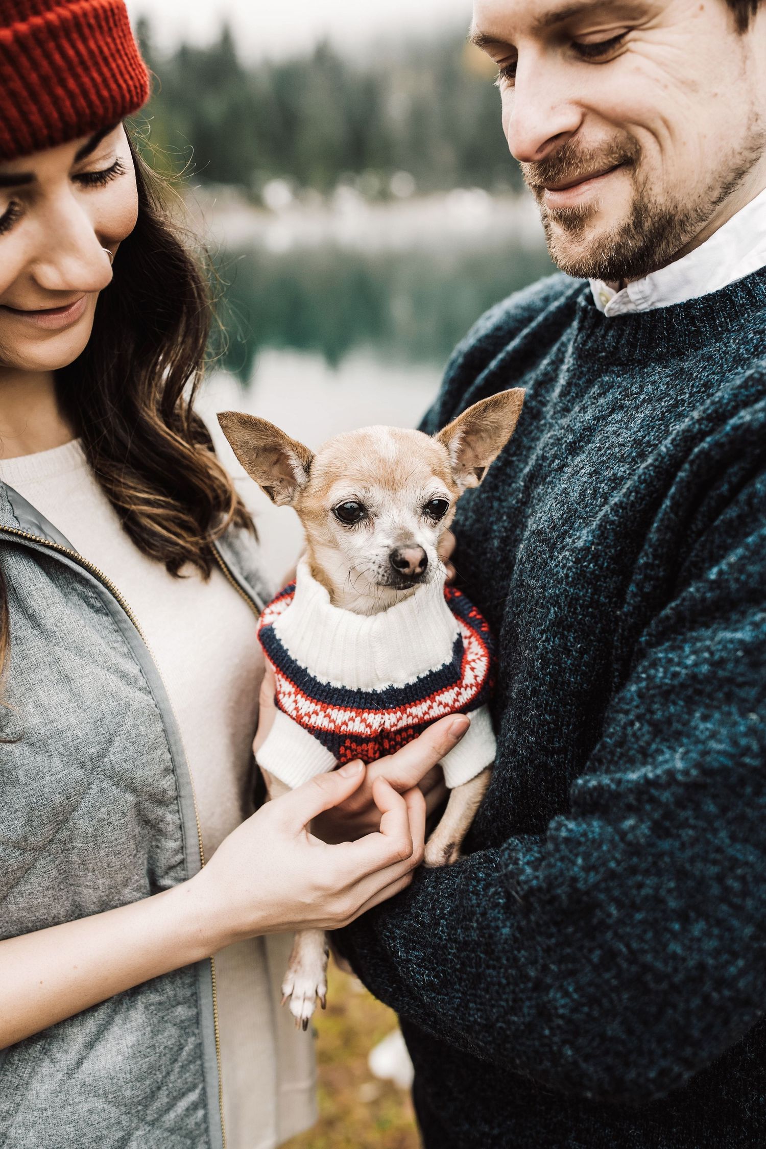 Couple holding a small Chihuahua in a knit sweater outdoors near a misty lake in autumn.