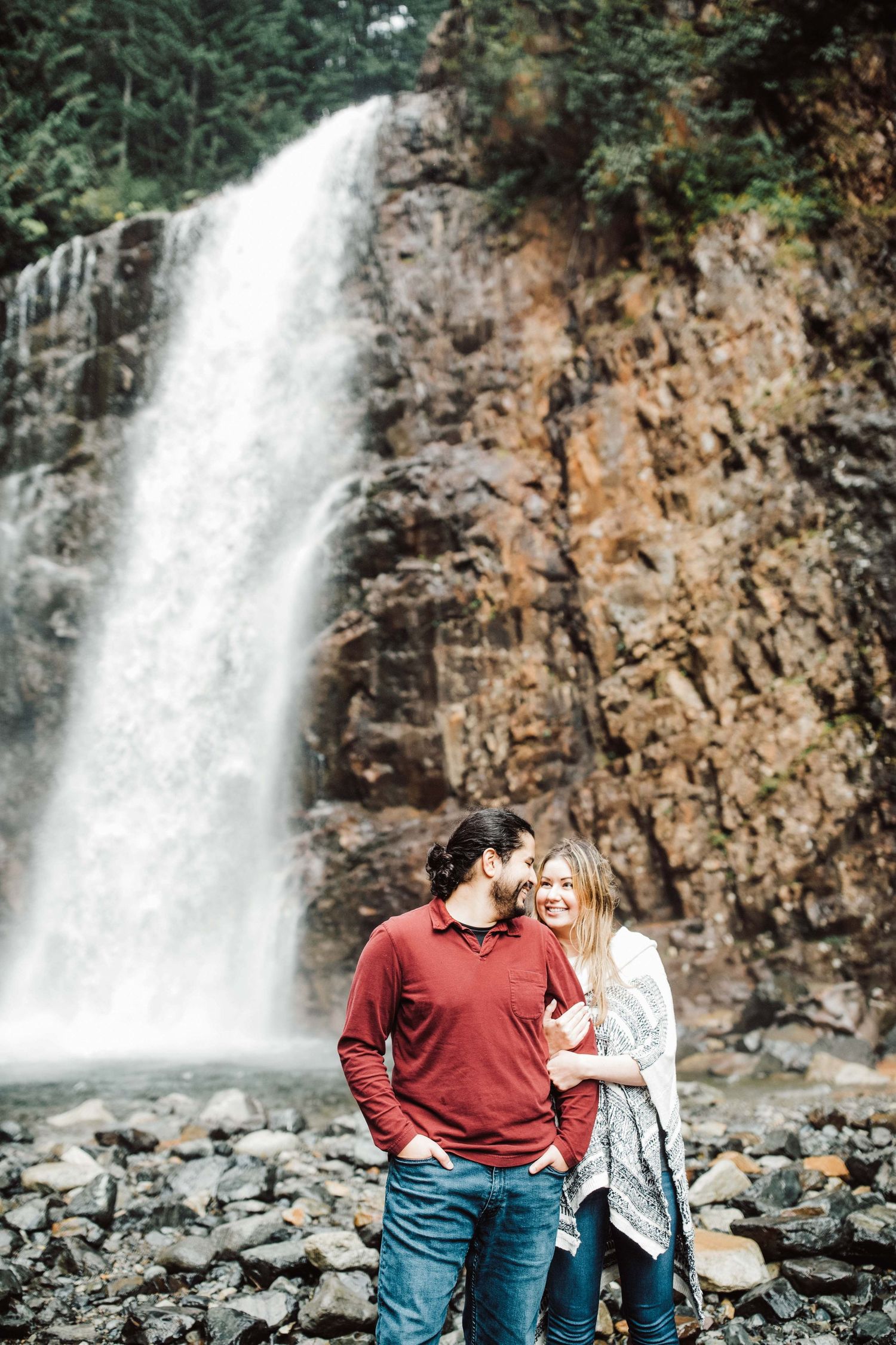 Couple embracing and smiling in front of a stunning waterfall cascading over rocky cliffs in a lush forest.