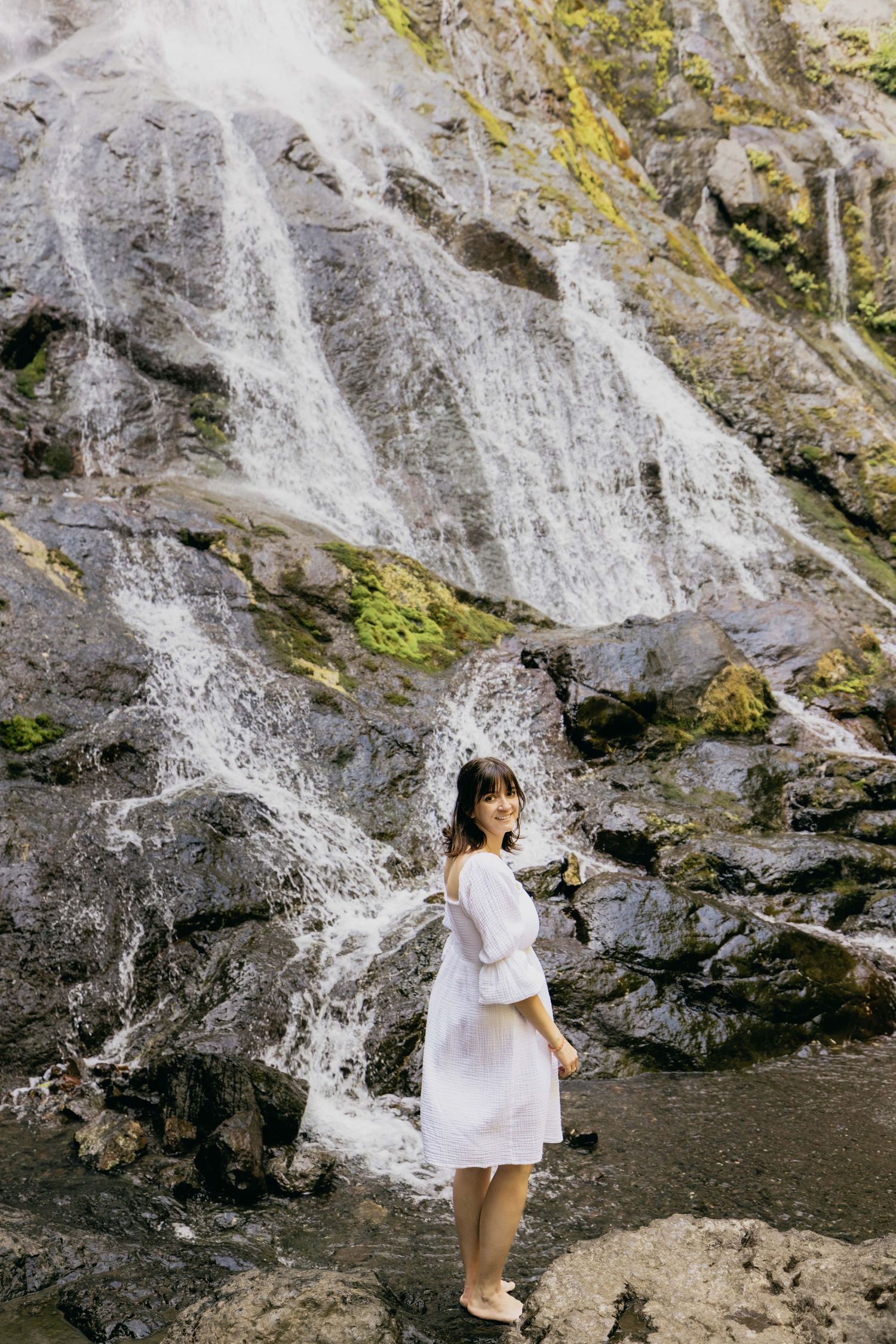 Woman in white dress standing before a cascading waterfall on rocky terrain.
