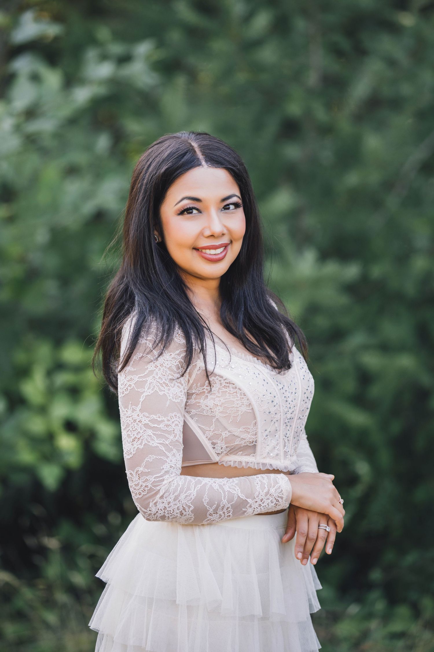 Portrait of a girl in a white lace dress smiles against a blurred green outdoor background.