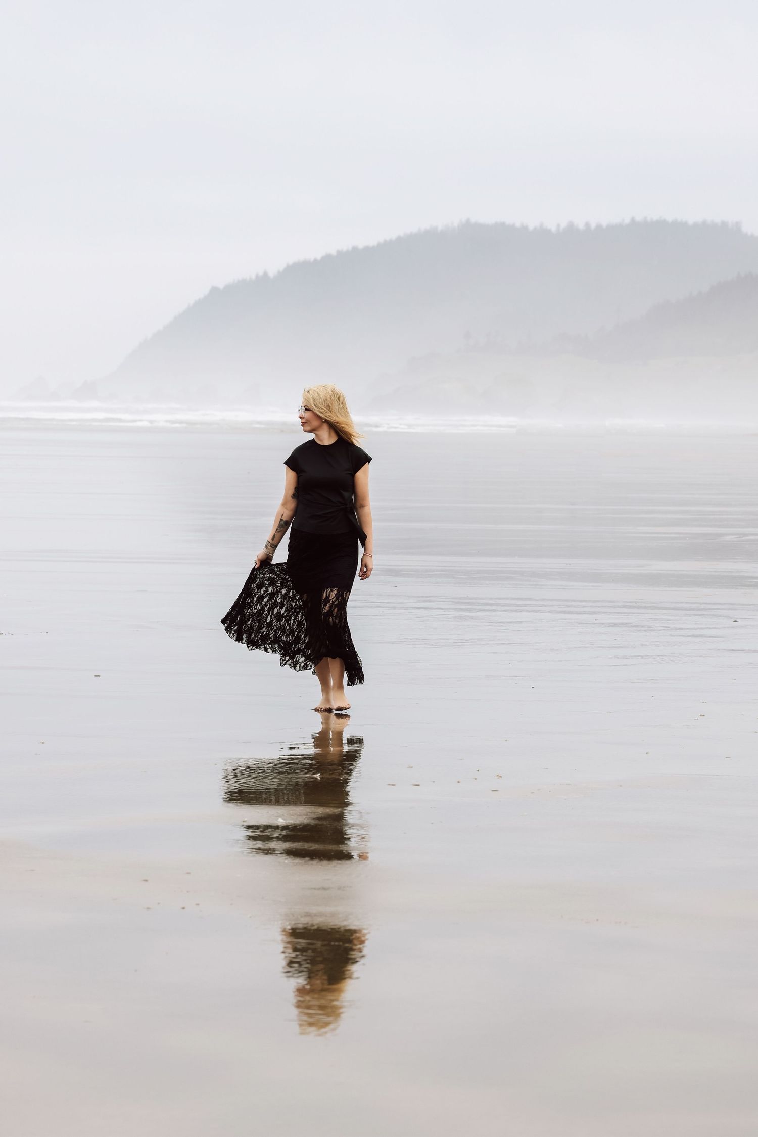Woman in black dress walking on misty beach with mountain backdrop, reflection visible on wet sand.