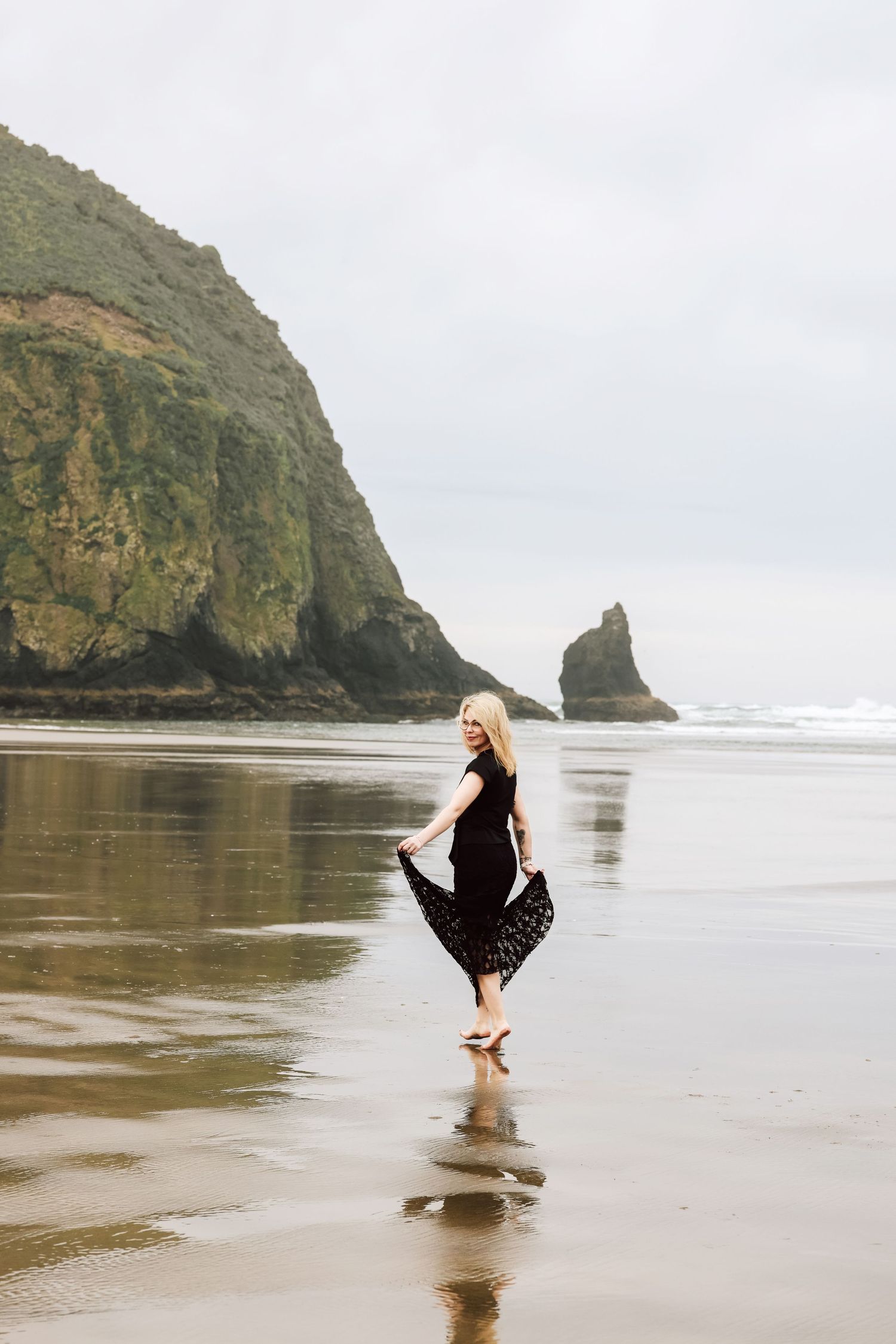 Blonde woman in black dress walking on reflective beach with dramatic rock formations and ocean backdrop.