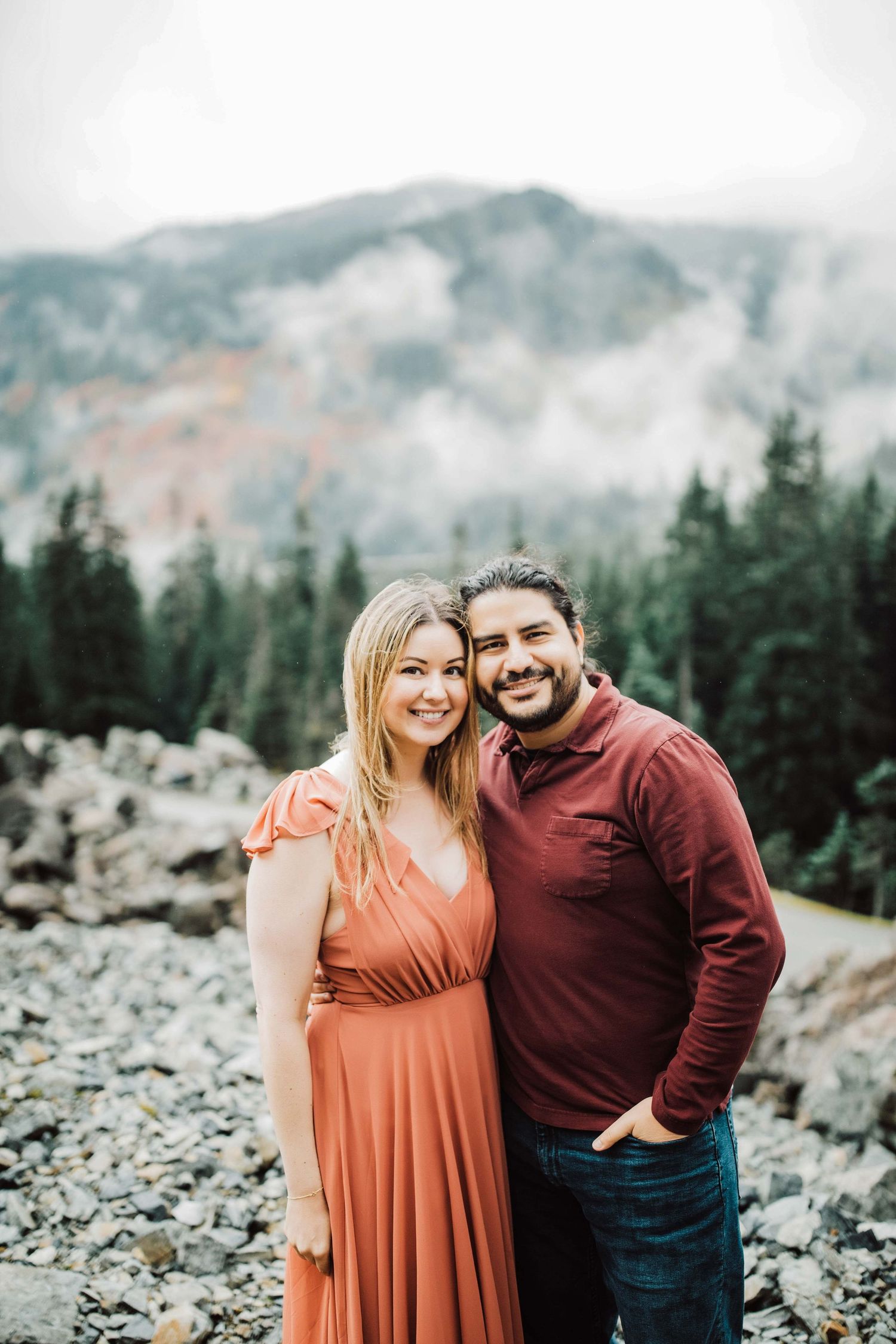 Couple smiling together in front of misty mountain forest backdrop, woman in coral dress, man in burgundy shirt.
