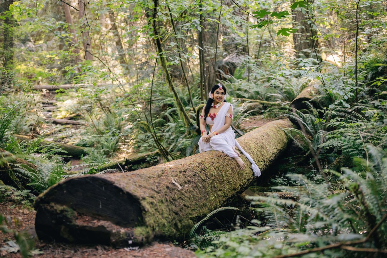 Woman in white dress sitting on a moss-covered fallen log surrounded by lush green ferns in a forest.