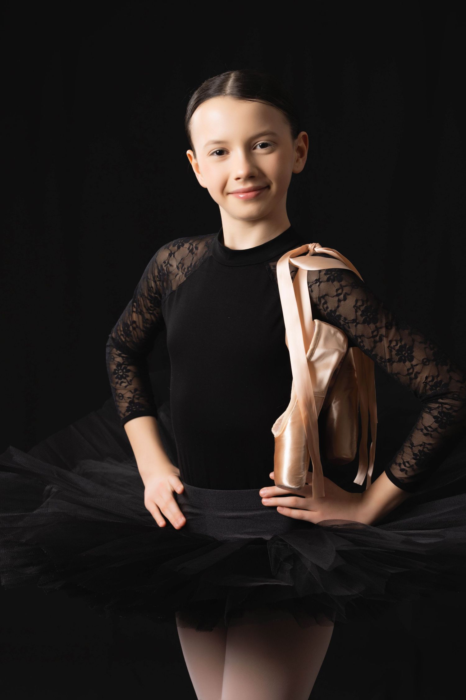 Young ballet dancer in black lace leotard and tutu poses gracefully against dark background in artistic portrait series.