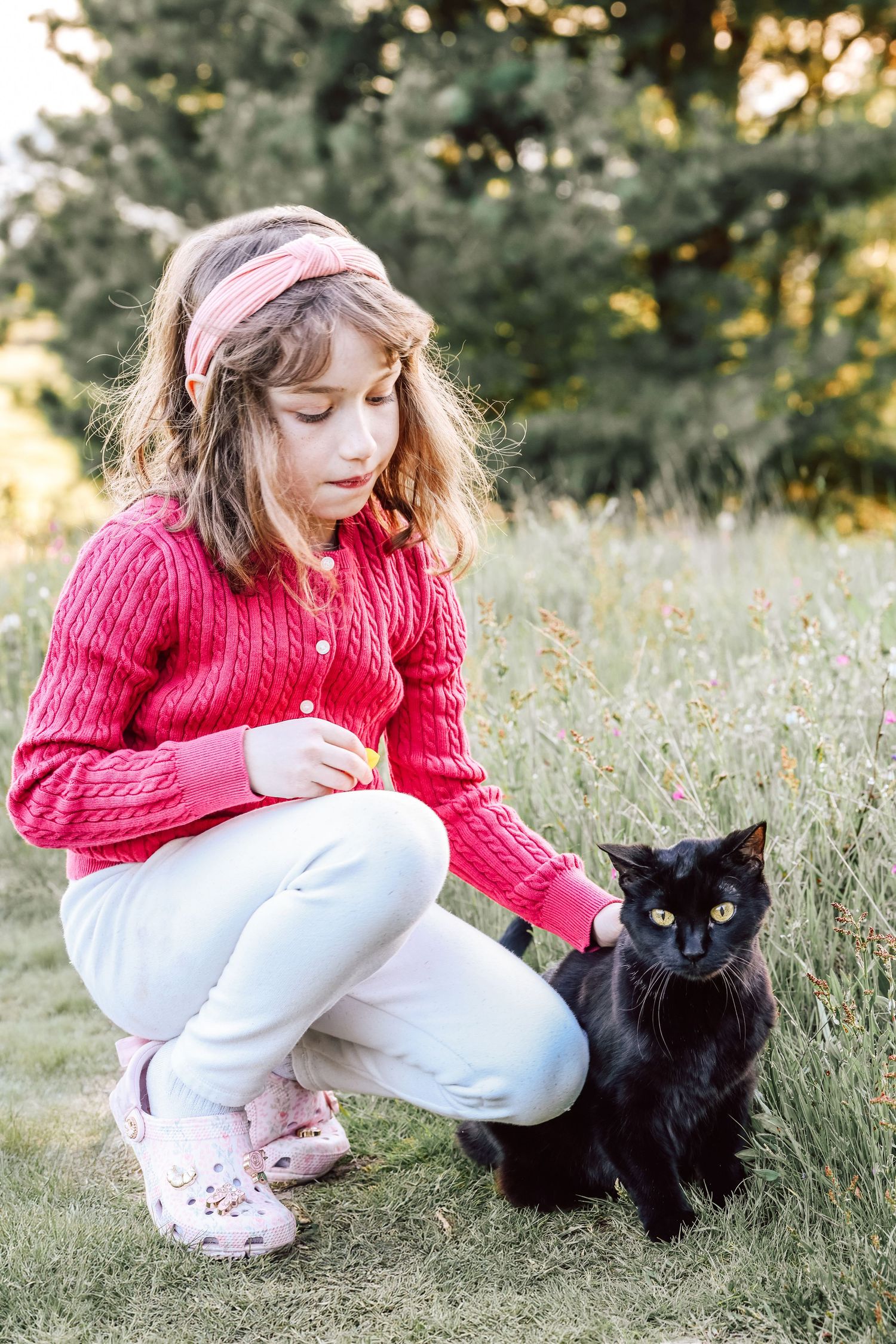 Young girl in pink sweater crouching outdoors, gently petting a black cat in a grassy field.