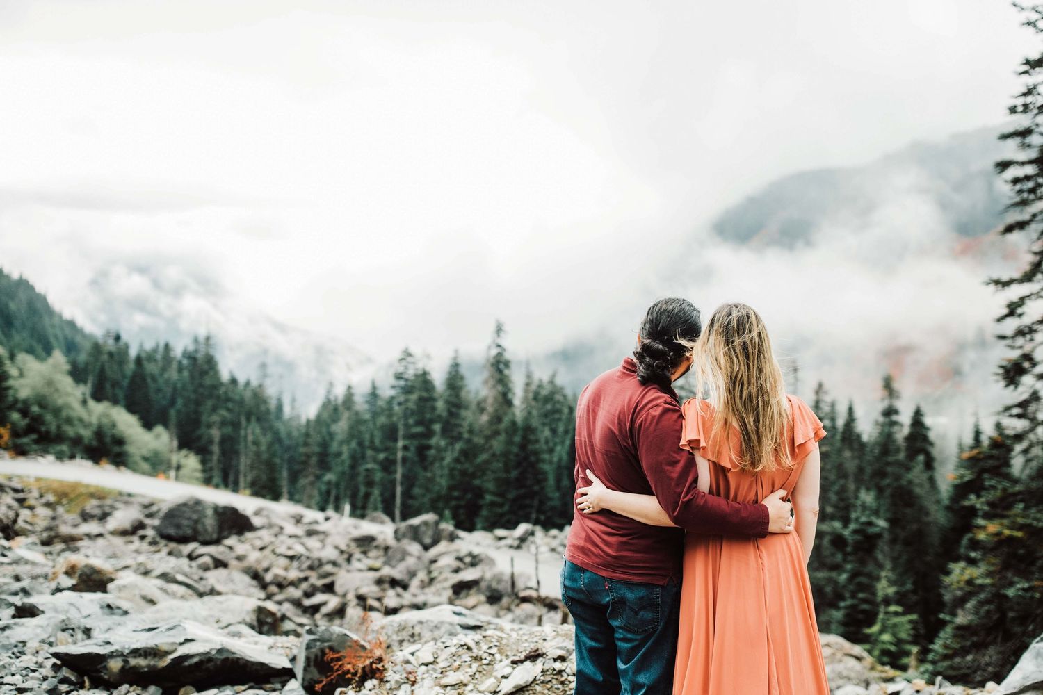Couple embracing while gazing at misty mountain forest landscape with rocky terrain and evergreen trees.