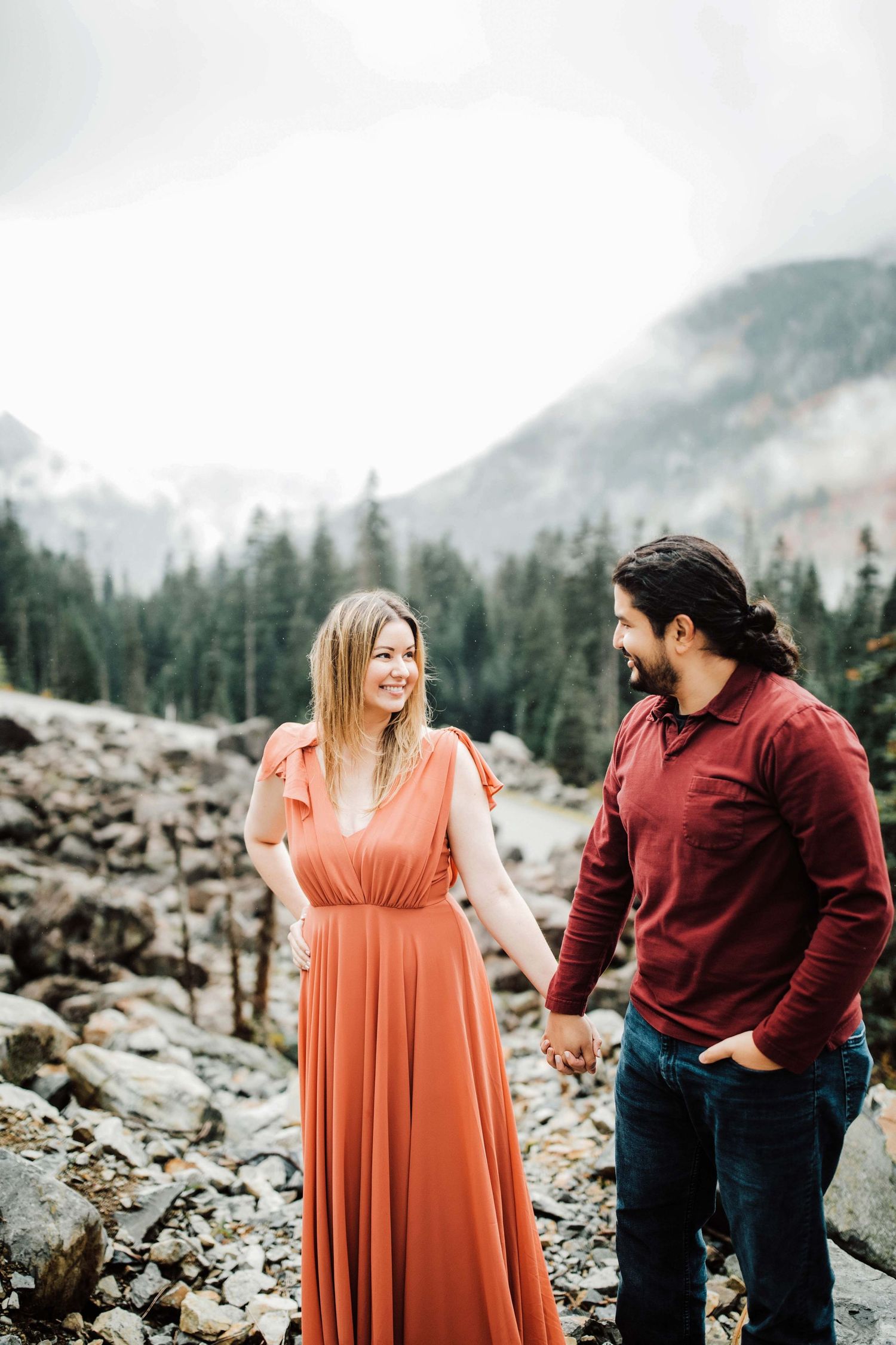 Couple holding hands smiling at each other in front of misty mountain forest, woman in coral dress, man in burgundy sweater.