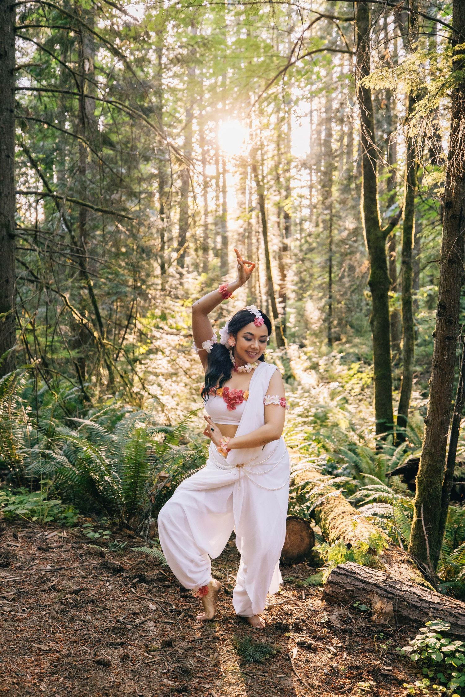 Woman in white traditional Indian attire performing classical dance in a sunlit forest setting.