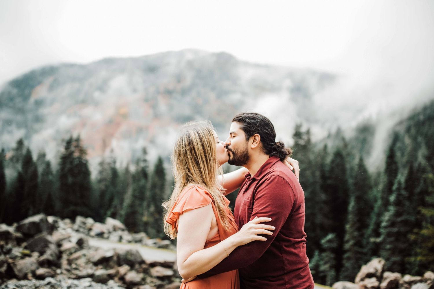 Couple sharing a romantic kiss outdoors with misty mountains and pine trees in the background.