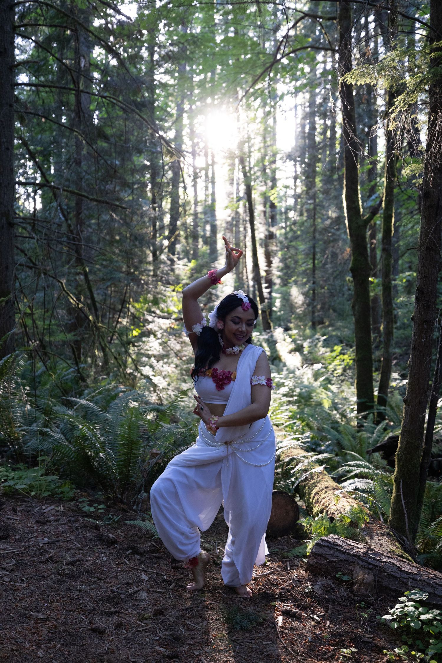 A woman in white traditional Indian attire performs a classical dance pose in a sunlit forest setting.