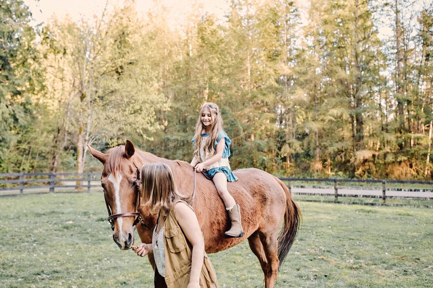 Young girl smiling while riding a chestnut horse in a green pasture surrounded by trees.
