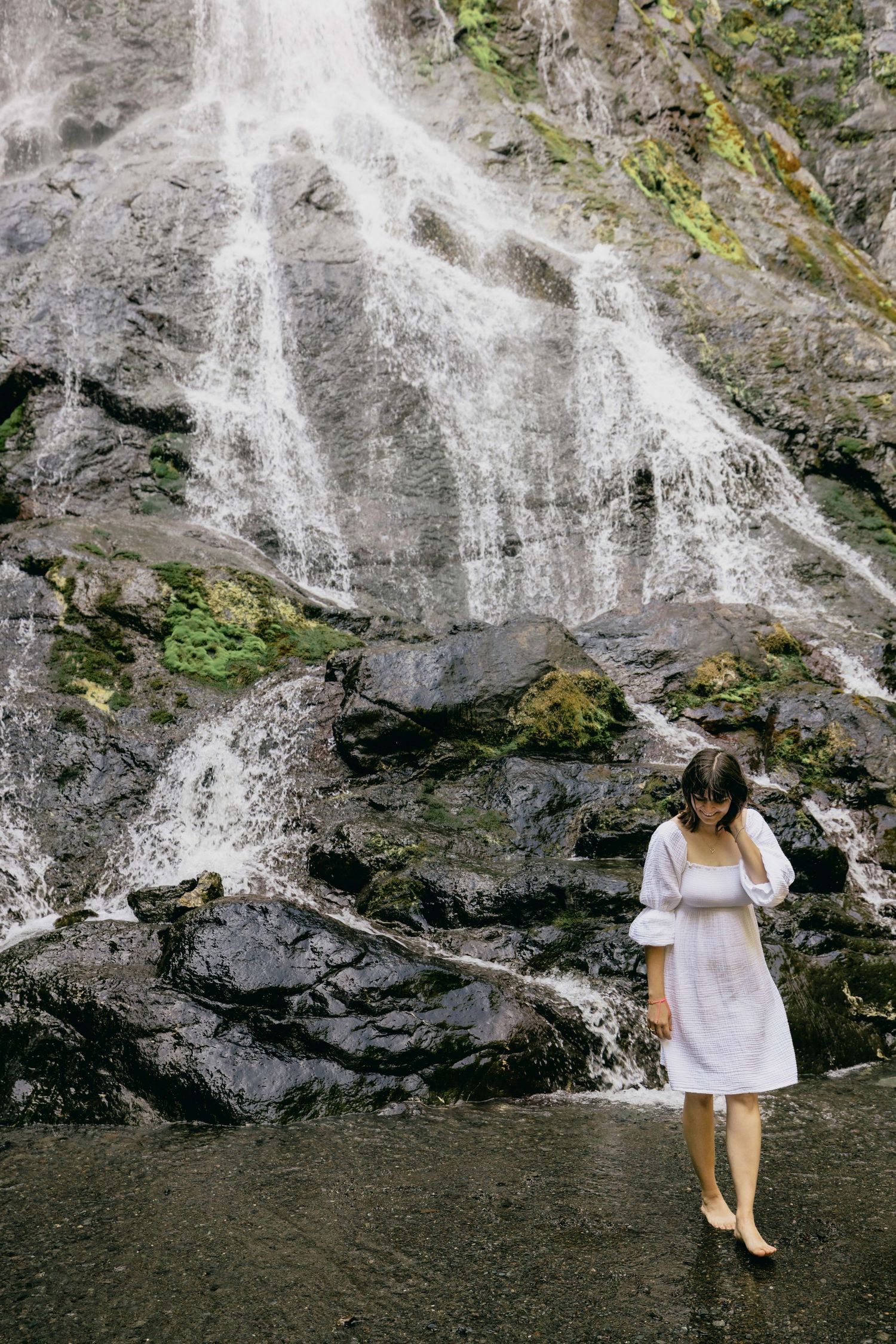 Woman in white dress standing barefoot on wet rocks before a stunning cascading waterfall surrounded by moss.