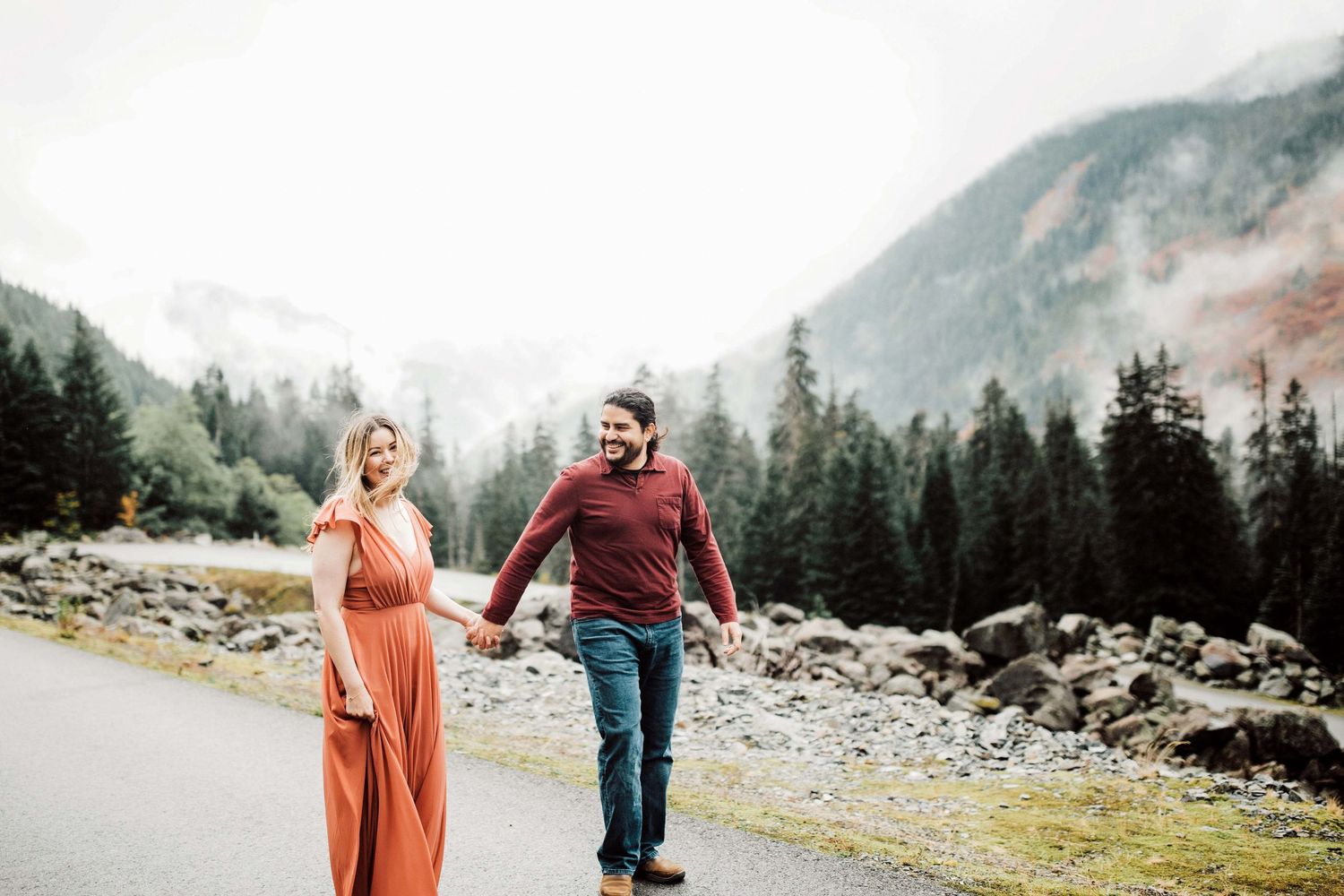 Couple holding hands walking on a misty mountain road surrounded by evergreen trees and foggy peaks.
