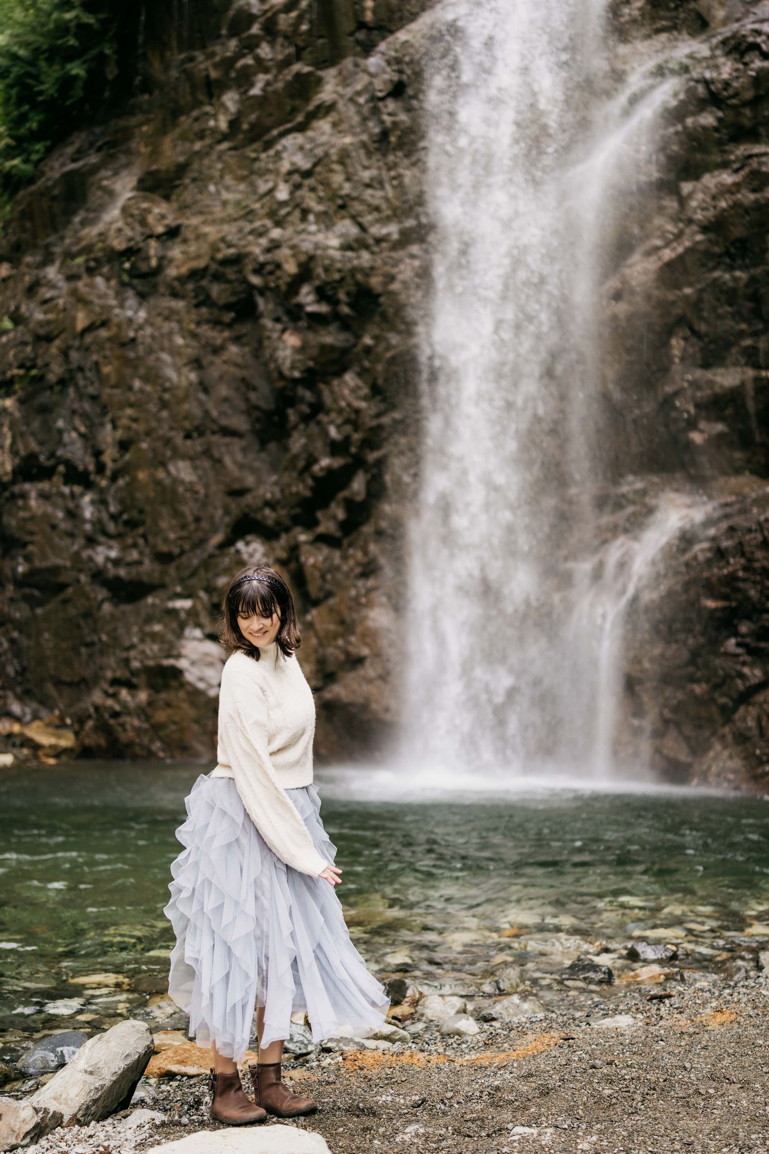 Woman in white sweater and blue skirt standing on rocks before a cascading waterfall in nature.