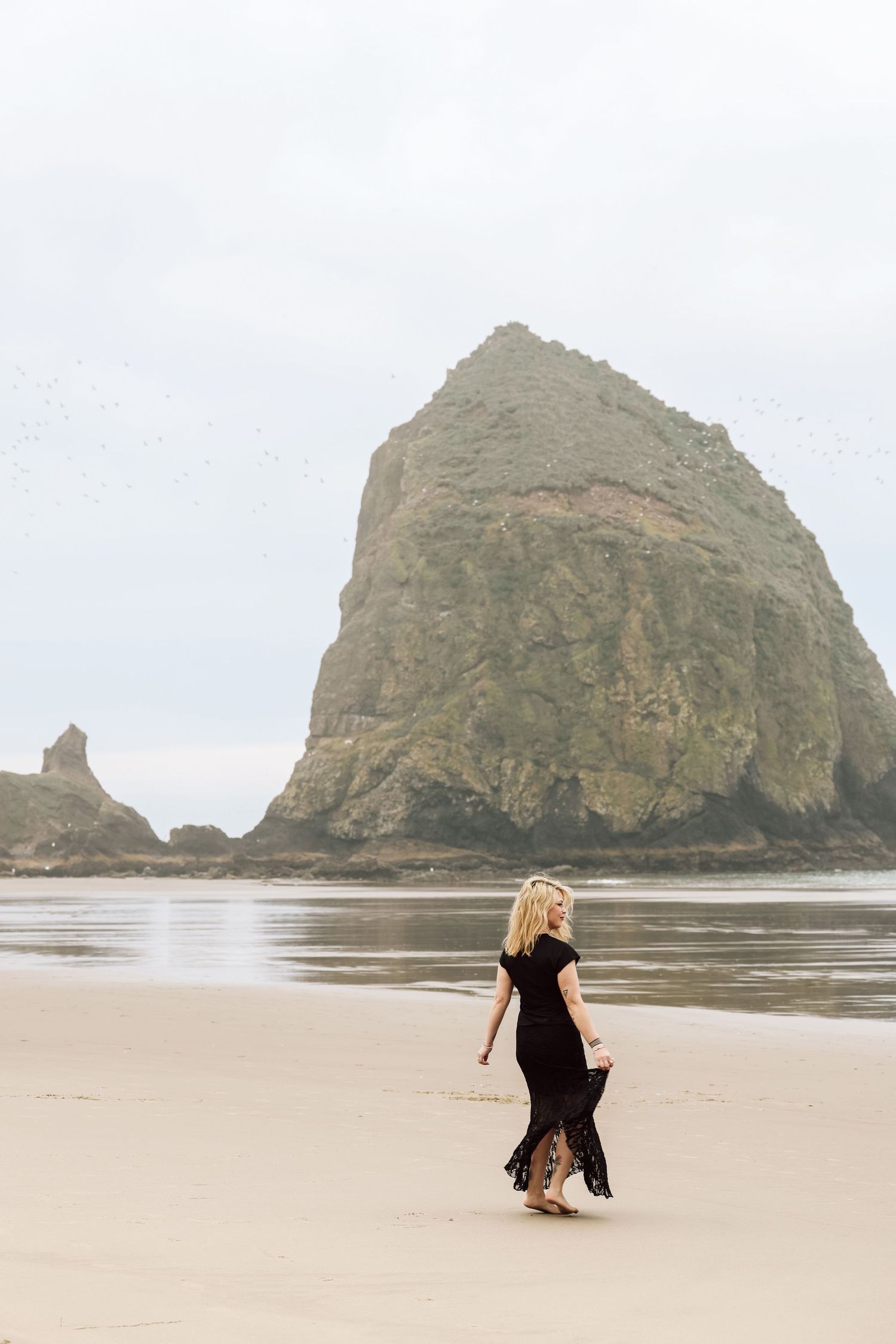 Blonde woman in black dress walking on sandy beach with iconic Haystack Rock at Cannon Beach, Oregon.