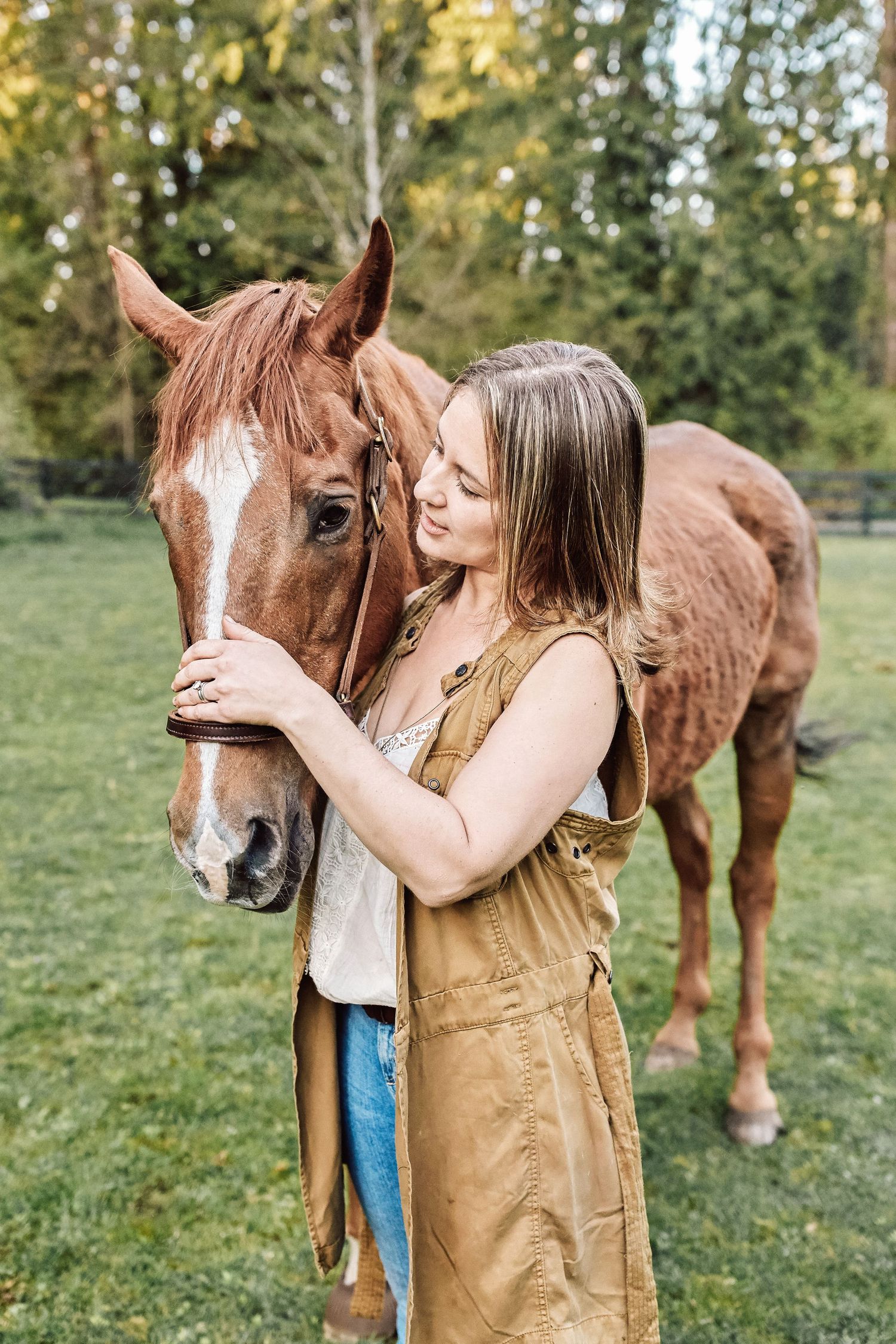 Young woman in overalls tenderly embracing a chestnut horse on a green pasture with trees in background.