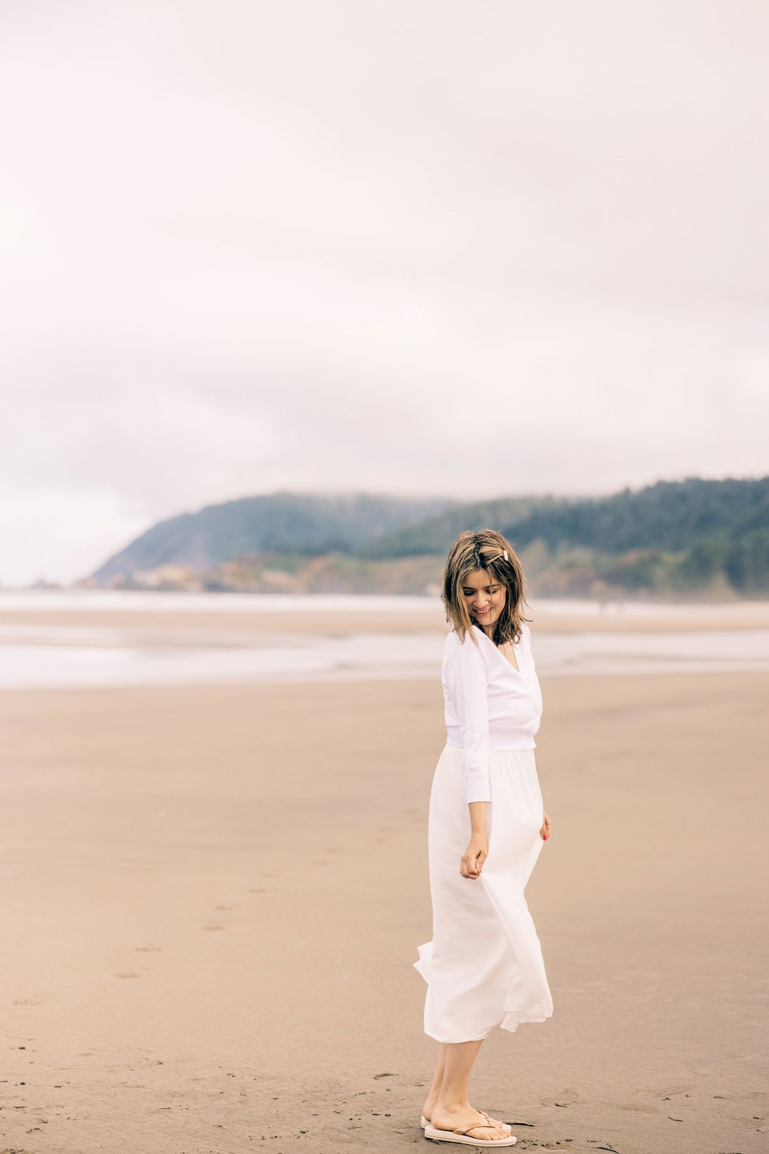 Woman in white dress walking barefoot on a sandy beach with misty hills in the background. Seattle adventure brand photographer for kids/pet