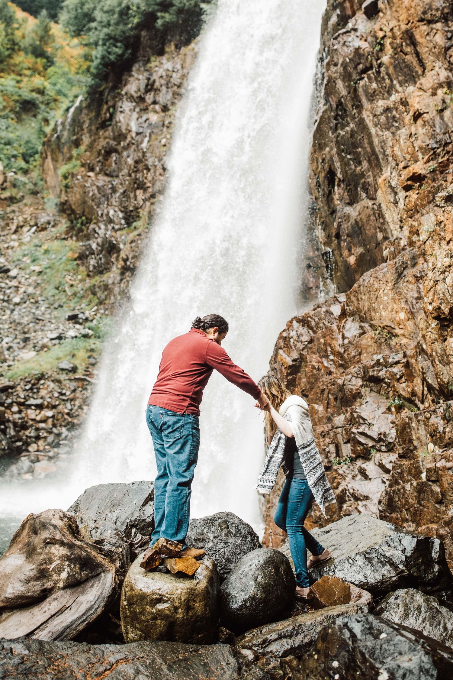 Couple holding hands while climbing rocks in front of a large waterfall in a scenic mountain landscape.