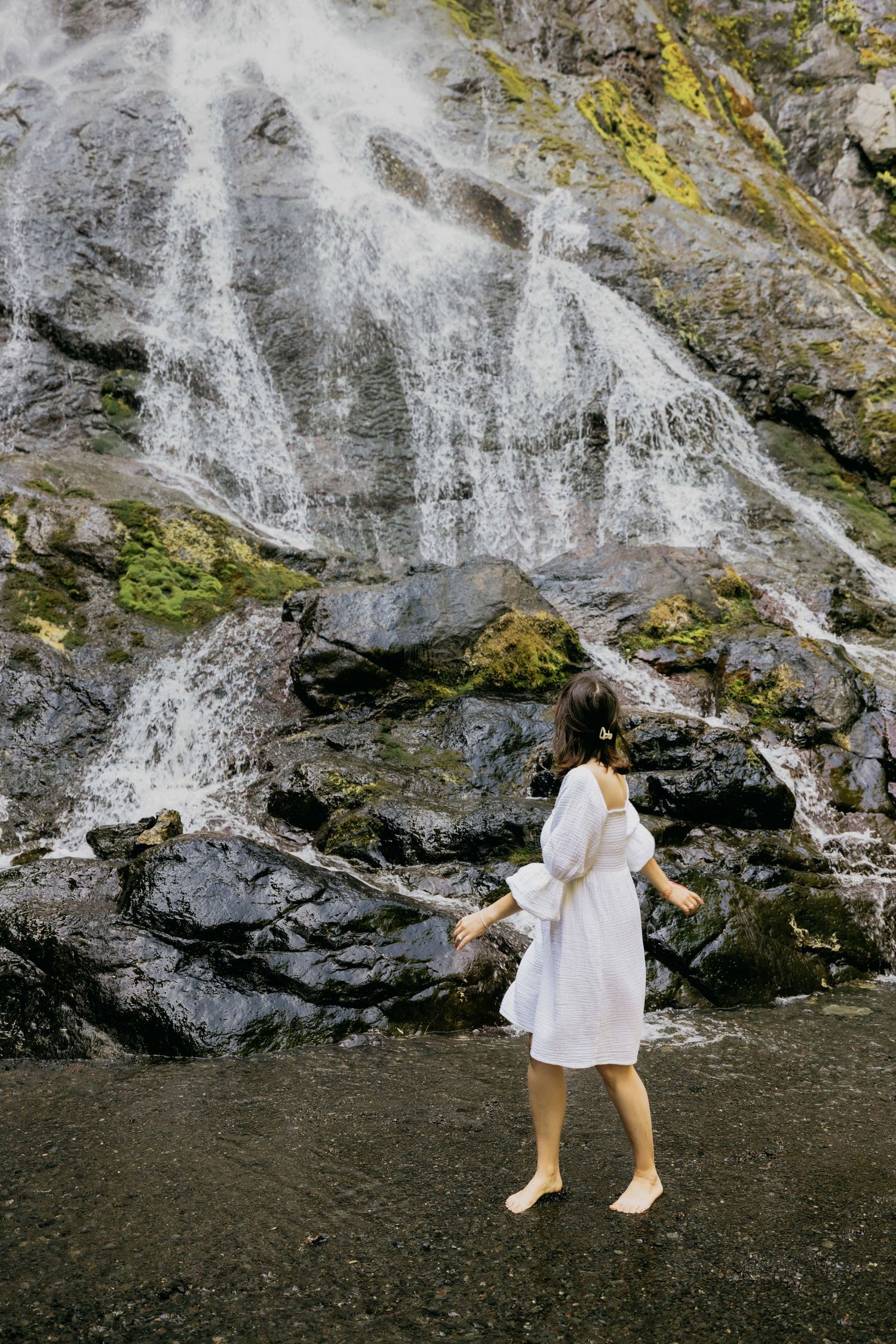 Woman in white dress standing barefoot near a stunning cascading waterfall on mossy rocks in nature.