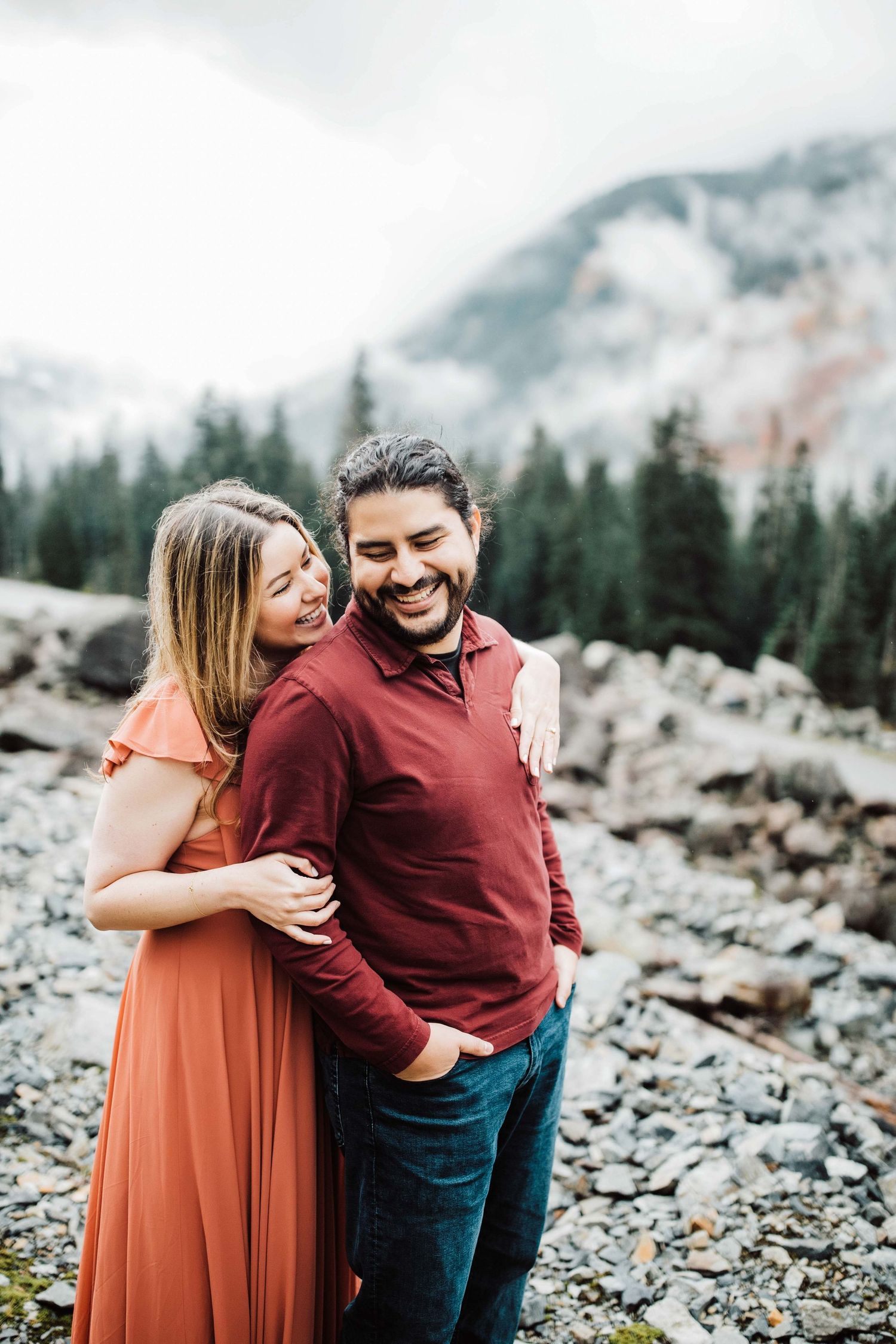 Couple laughing and embracing outdoors on rocky terrain with snow-capped mountains and evergreen trees in background.