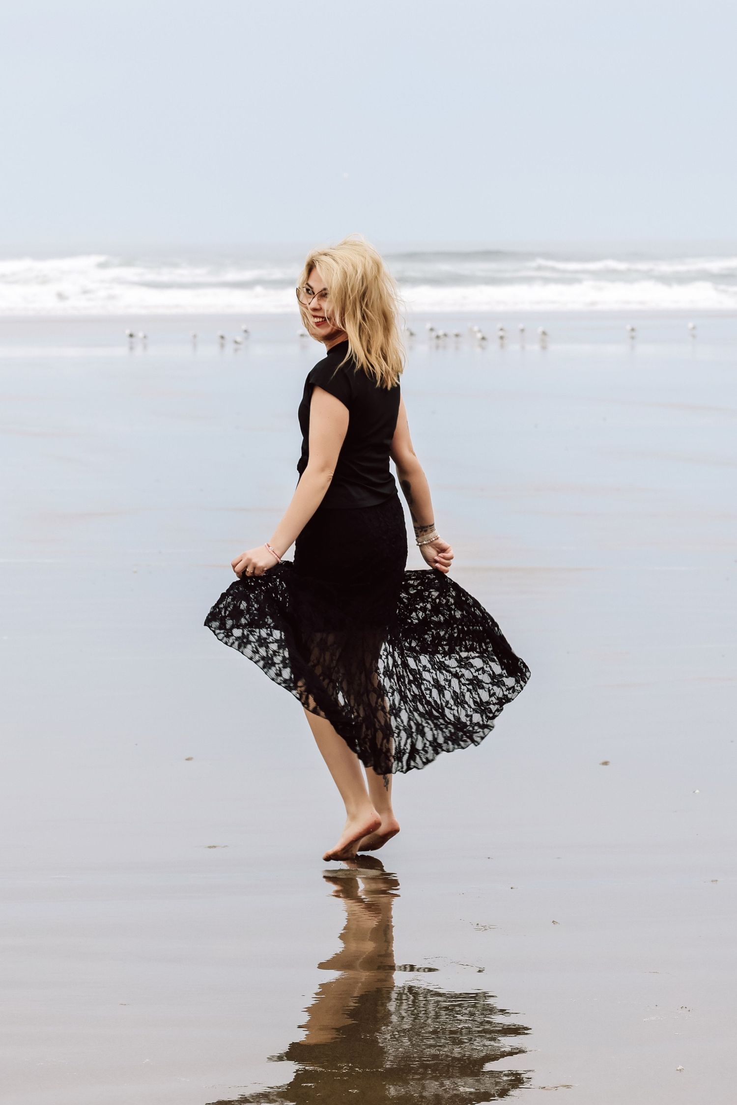 Blonde woman in black lace dress walking barefoot on a sandy beach with ocean waves in the background.