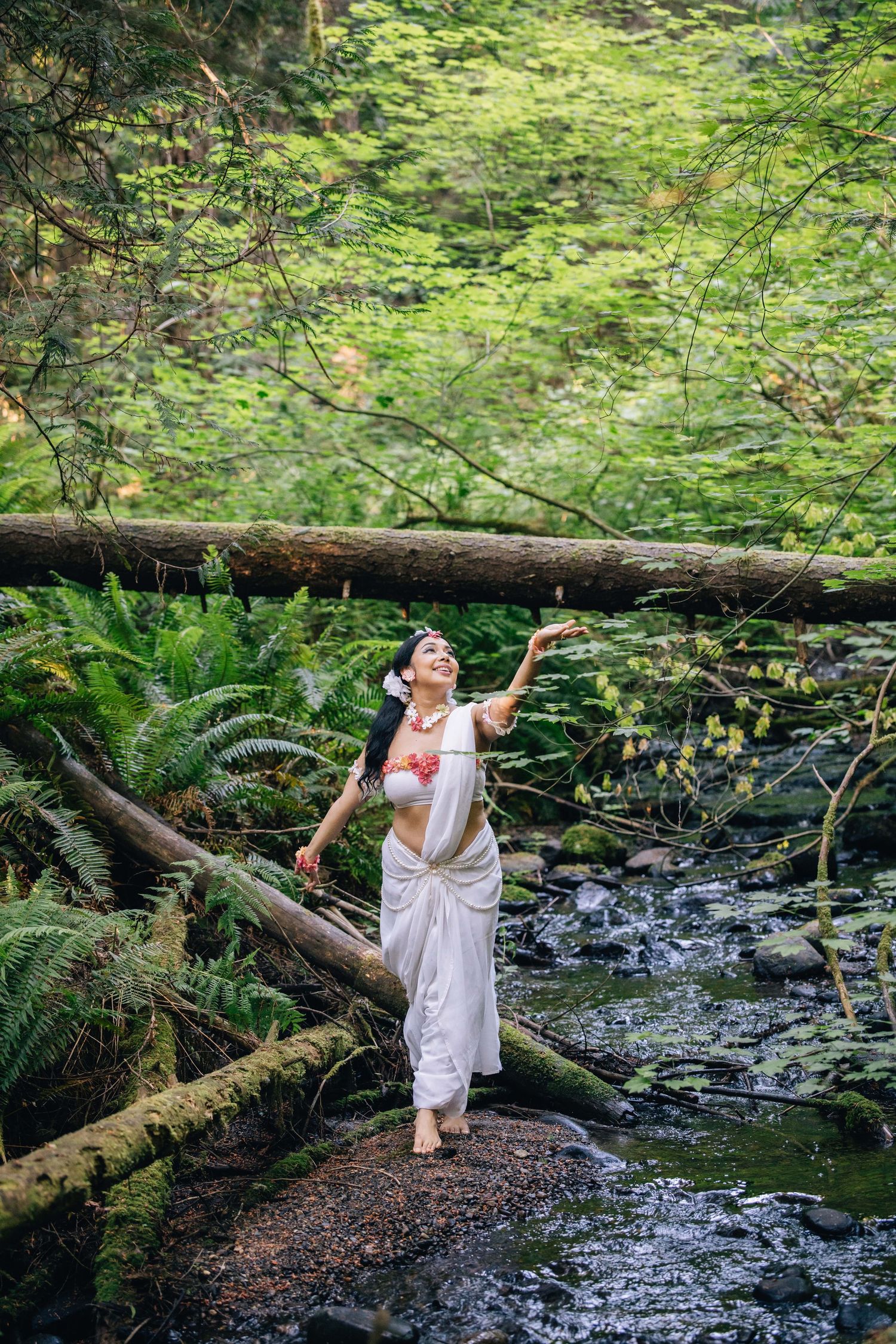 Woman in white sarong and floral top walks barefoot on mossy log over forest stream surrounded by lush green ferns.
