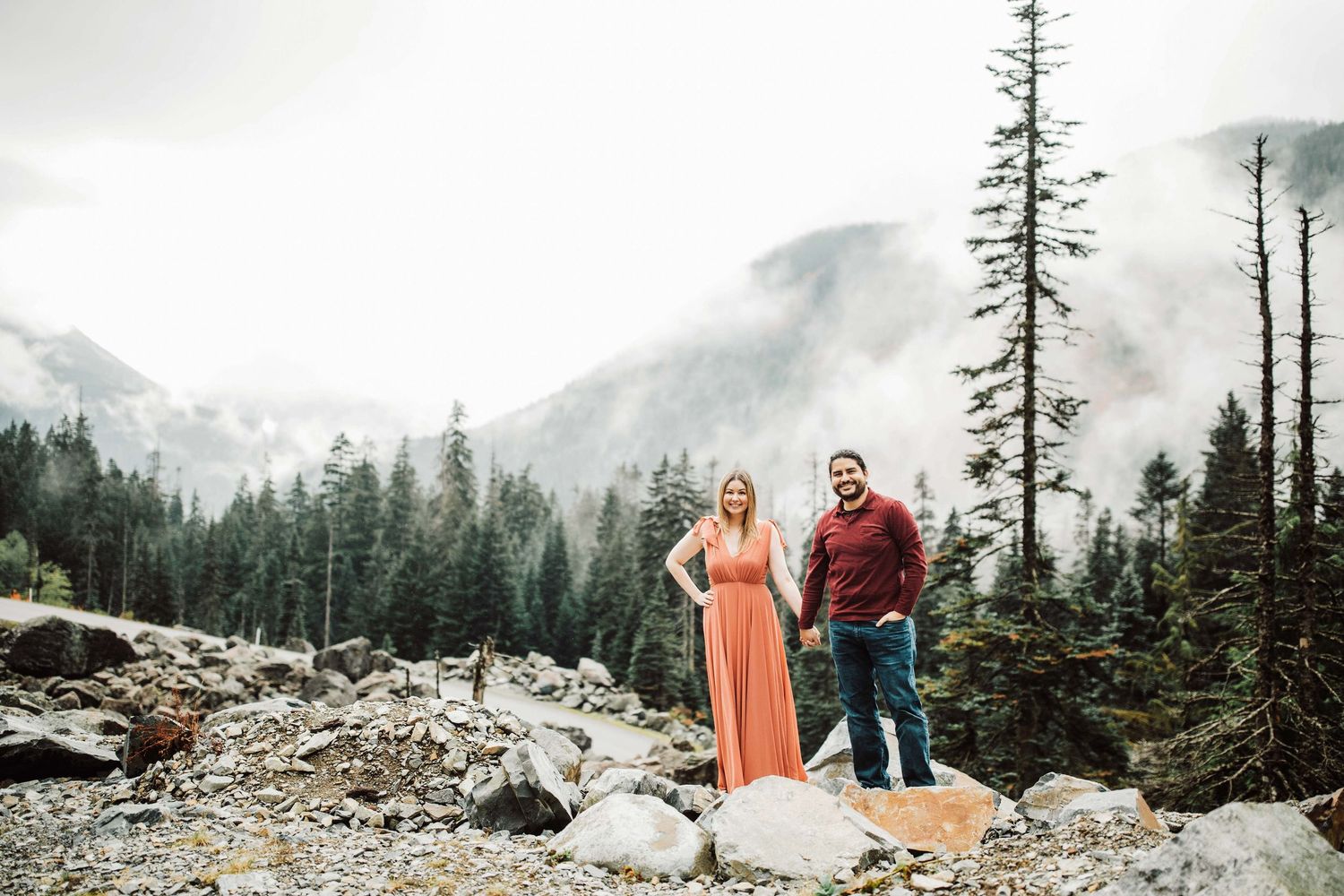 Couple holding hands in a rocky mountain forest with misty peaks, woman in coral dress, man in jeans.