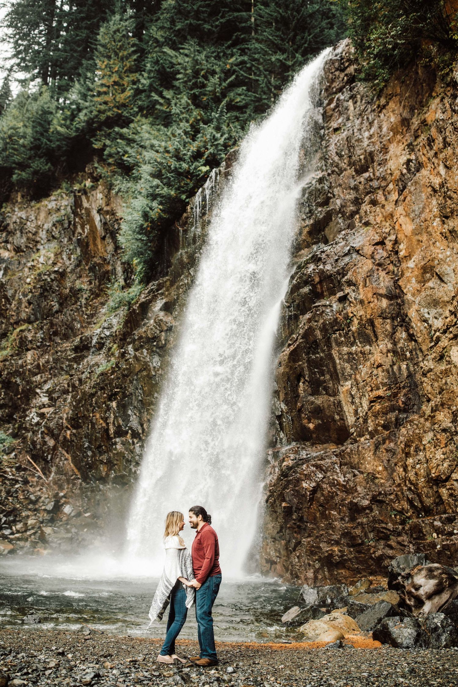 Couple standing romantically in front of a stunning waterfall surrounded by rocky cliffs and lush evergreen trees.