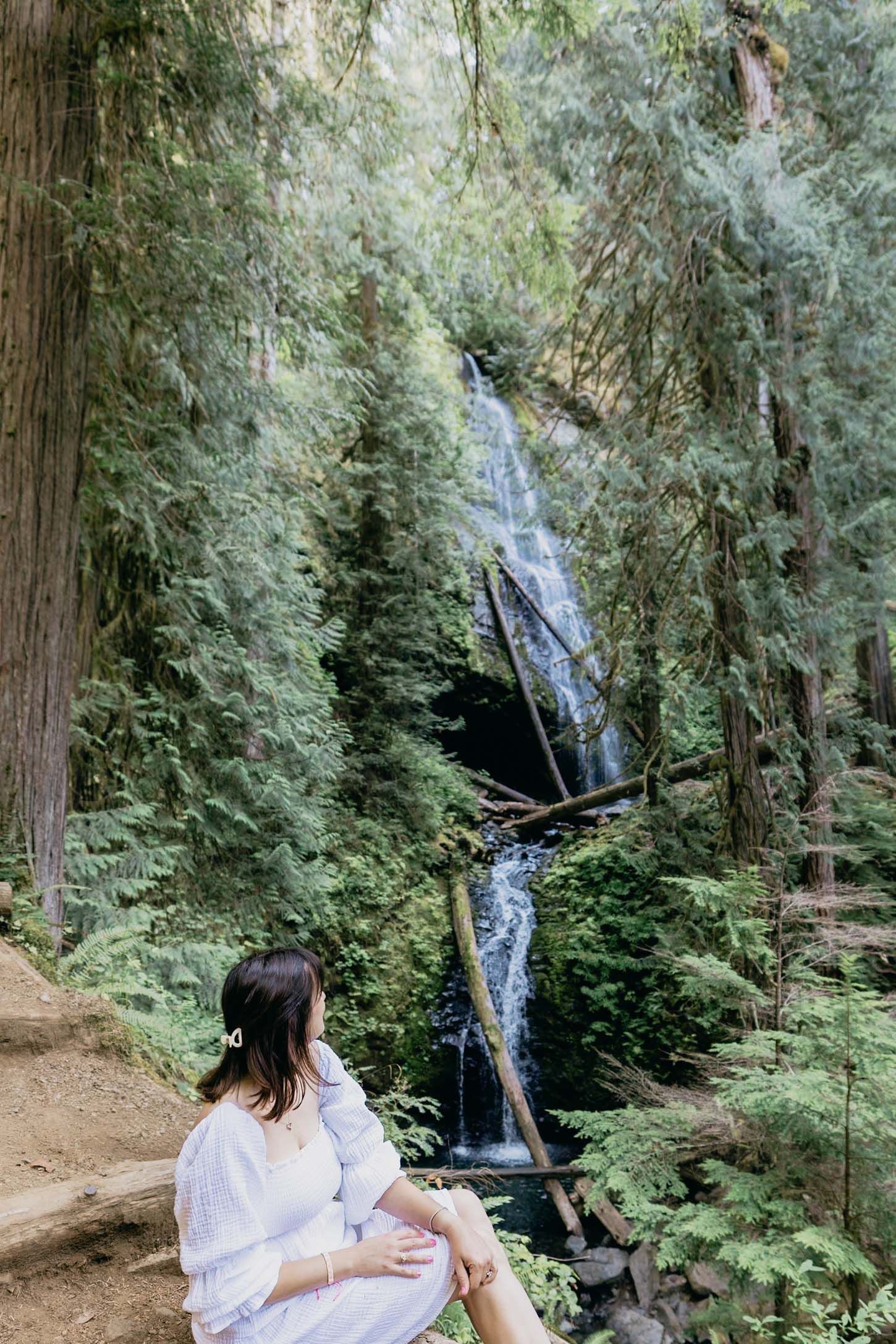 Woman in white dress sitting near a serene waterfall surrounded by lush green forest trees.