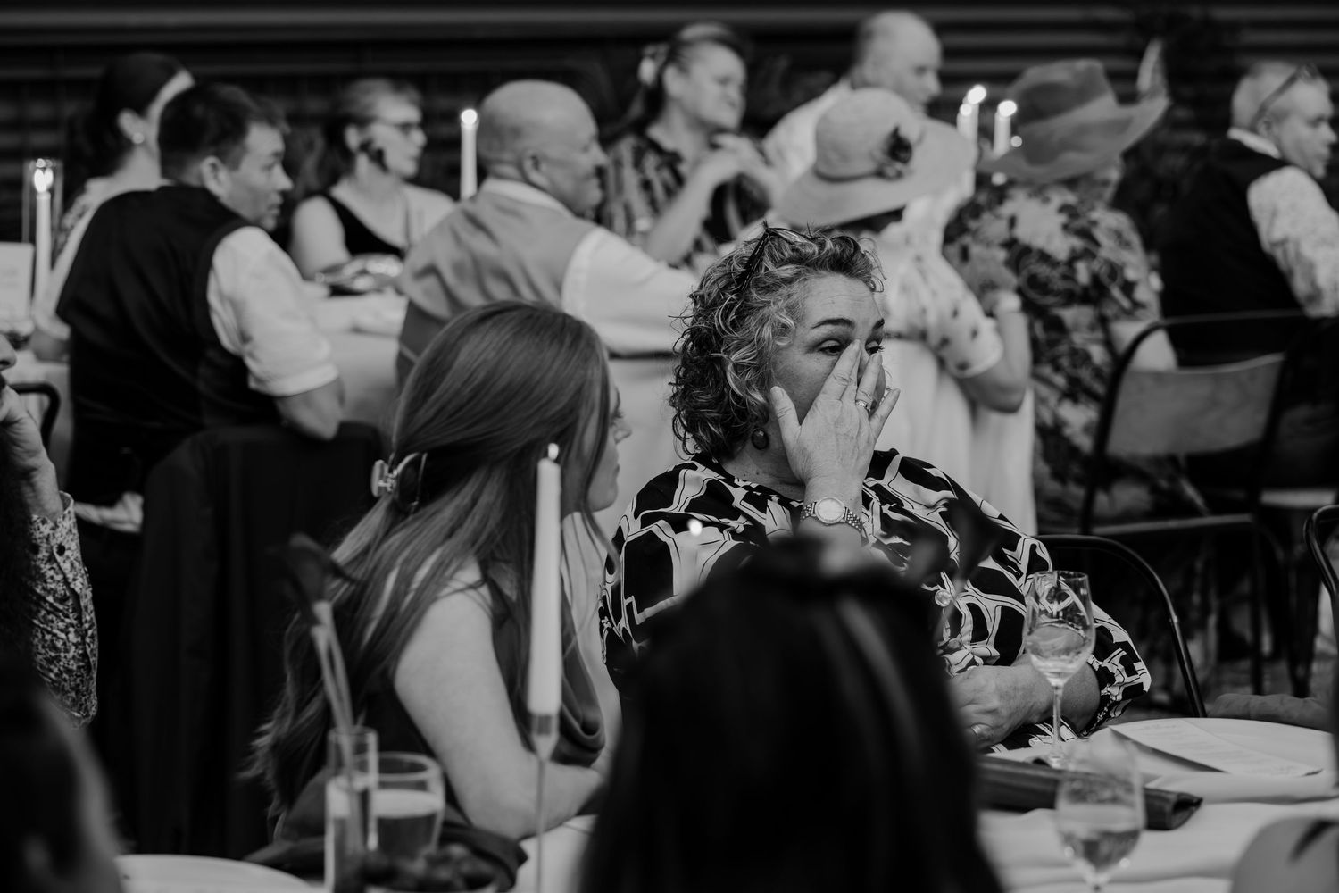 Black and white photo of guests seated at tables during a formal dinner event, with a woman covering her mouth.