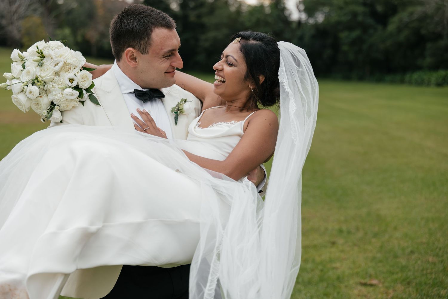 Groom carrying bride in white wedding dress and veil on green lawn, both smiling at each other.