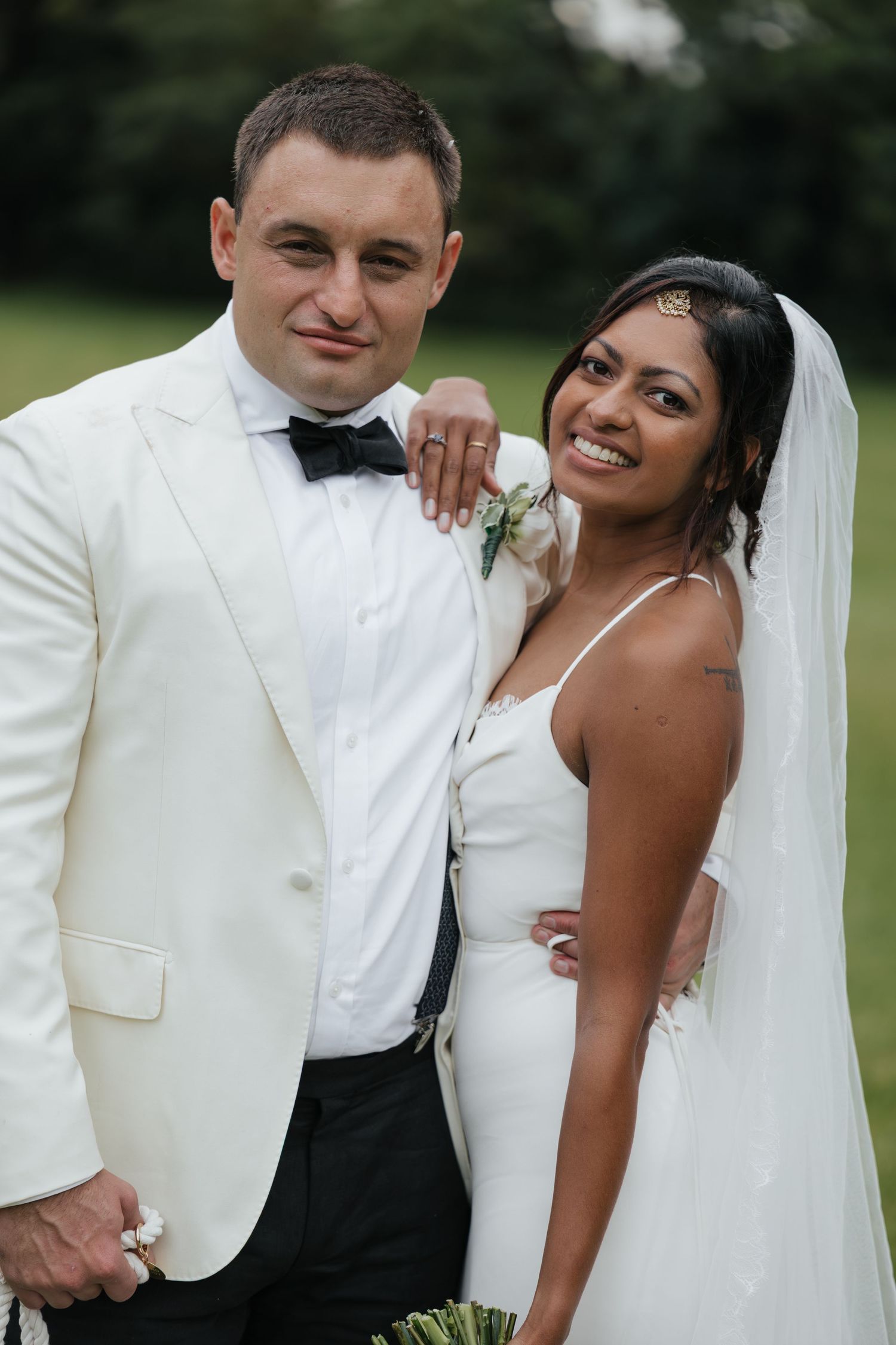 Interracial couple posing in wedding attire outdoors, groom in white tuxedo, bride in white gown with veil.