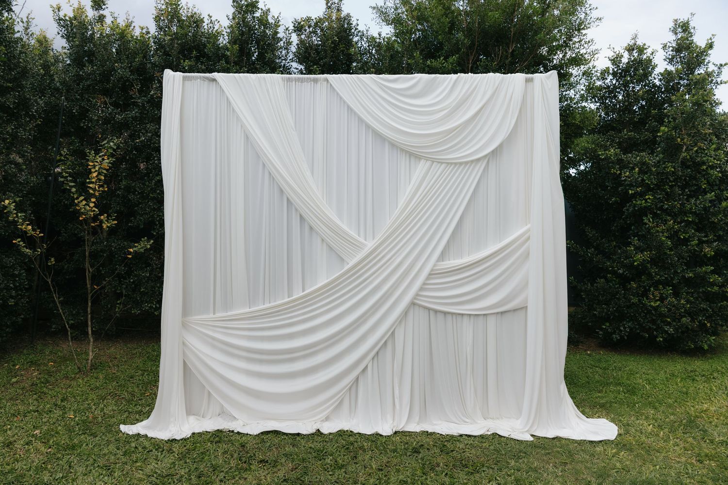 Elegant white draped fabric backdrop with crossed swags, perfect for weddings or events, set outdoors on green lawn.
