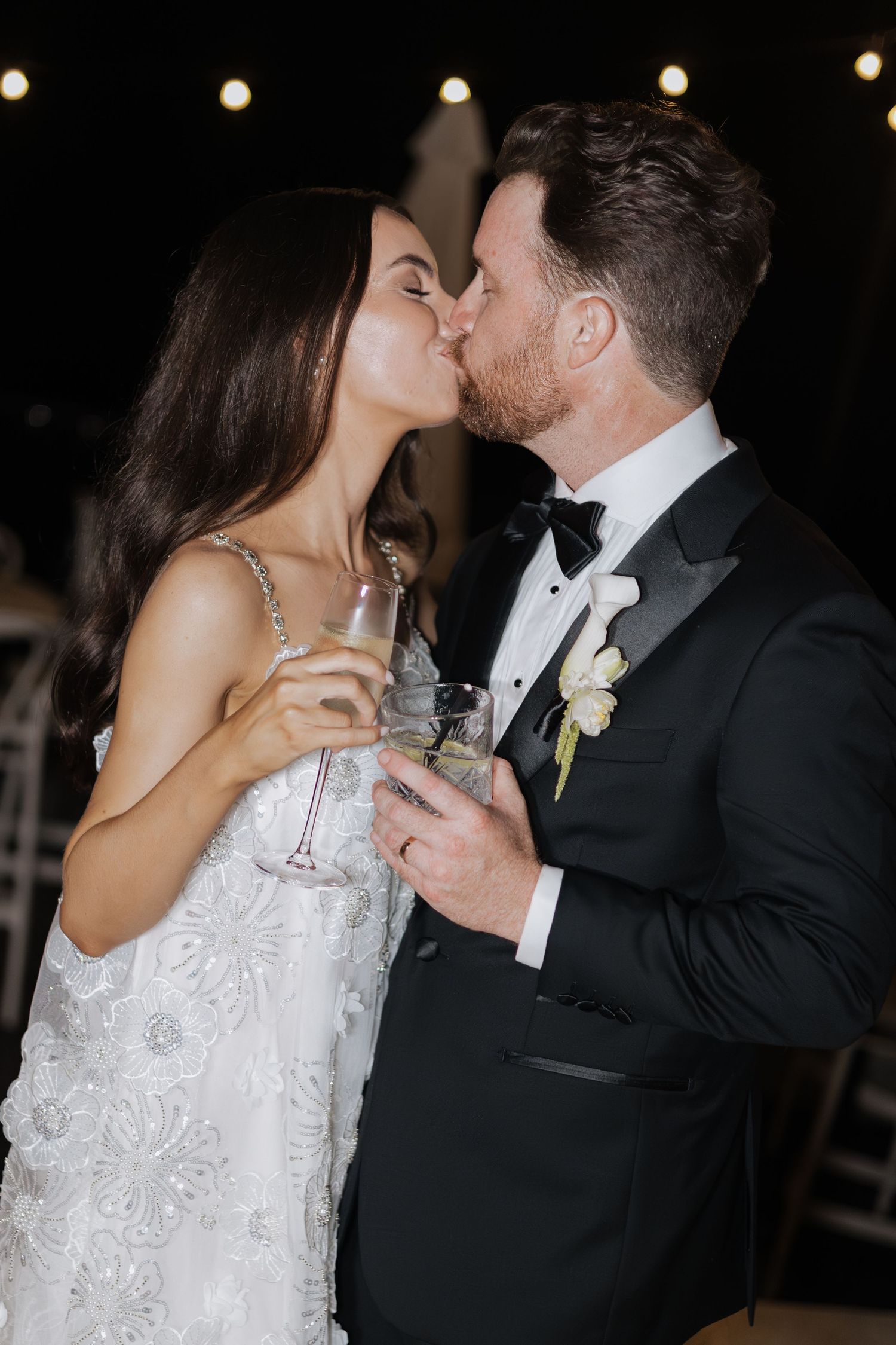 Bride and groom share a kiss while holding champagne glasses at their elegant wedding reception.