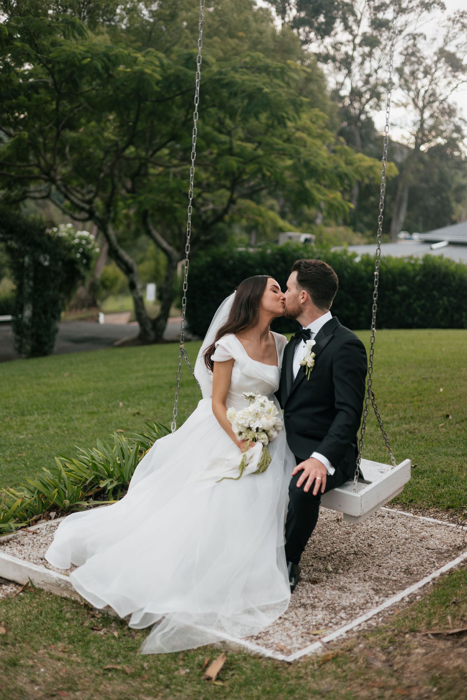 Bride and groom kissing on a wooden swing in a lush garden, bride holding white bouquet in elegant wedding attire.