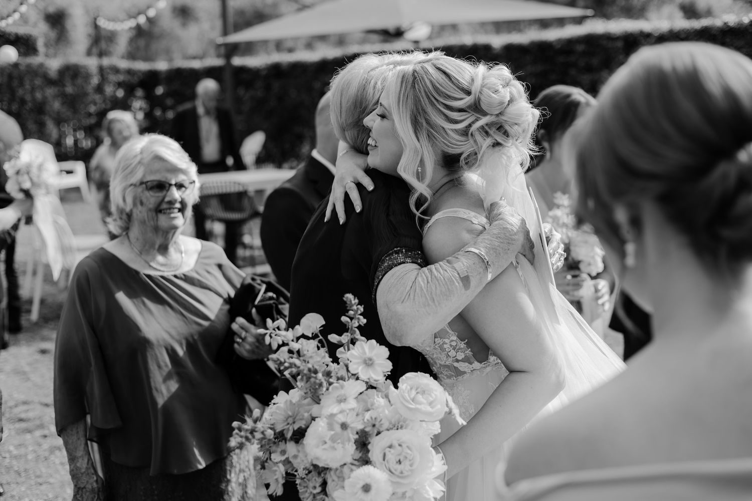 Black and white wedding photo of bride embracing guest while older woman smiles nearby, holding floral bouquet.